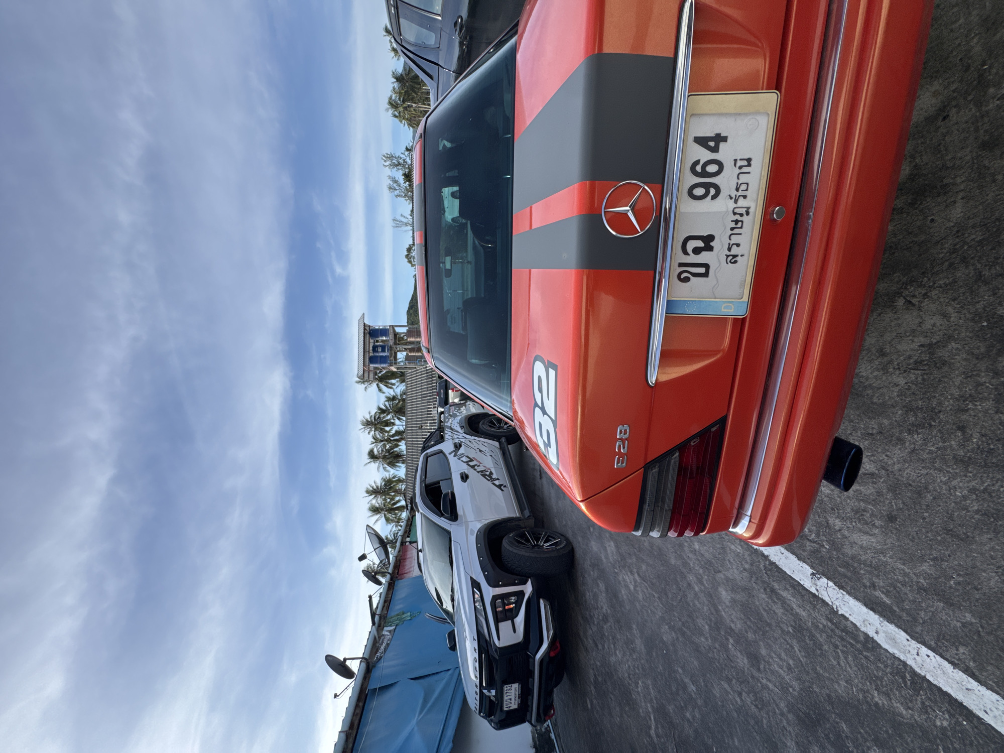 The image depicts an outdoor parking area in ตำบลเกาะพะงัน, Thailand, likely a resort or residential complex, with a bright, partly cloudy sky. In the foreground, the rear of a bright red Mercedes-Benz E28 is prominently featured, showing its taillight, bumper, exhaust pipe, and license plate which reads "ขจ 964 สุราษฎร์ธานี". The car has a racing stripe design with two dark gray stripes down the center of the trunk, and a "32" graphic on the trunk lid. To the left of the Mercedes, a white Mitsubishi Triton pickup truck is parked, displaying its front and side profile. The truck has a "TRITON SPORT" decal on the side and a license plate that reads "602 1792". In the background, a cluster of palm trees and some buildings, including what appears to be a lifeguard tower or small elevated structure, are visible. Satellite dishes are mounted on the roof of a blue-covered structure behind the pickup truck. The ground is paved with asphalt and marked with a white line.