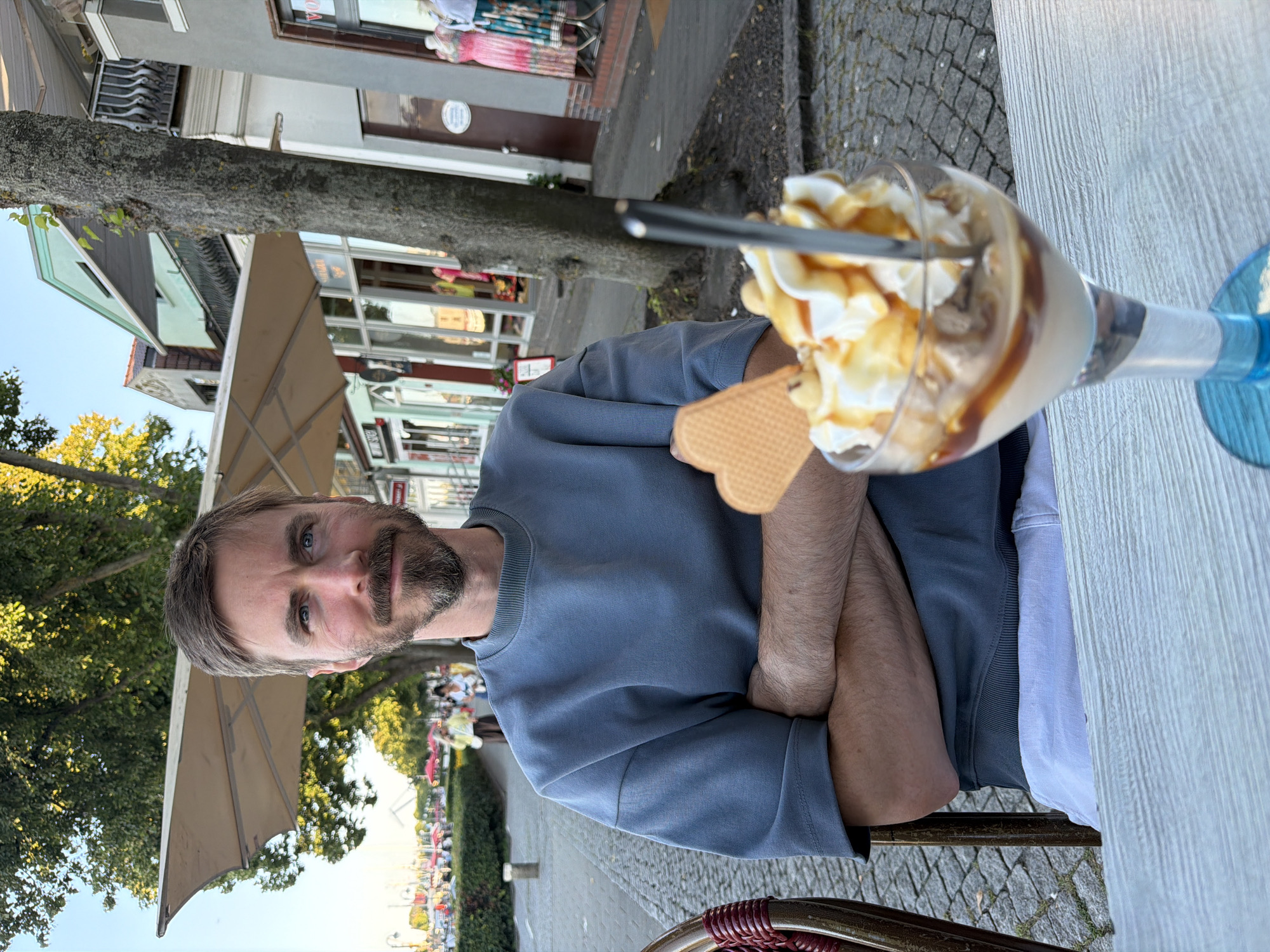 A man sits at a table with his arms crossed, looking at the camera. He has a beard and mustache and is wearing a blue sweatshirt. In front of him is a dessert in a glass, topped with whipped cream and caramel sauce, with a waffle on the side. The setting appears to be an outdoor cafe. The man is situated in front of a cobblestone sidewalk and a row of buildings. In the background, there are trees and people walking. This location is consistent with the promenade along the Warnow River in Rostock, Germany, a popular area with cafes and shops.