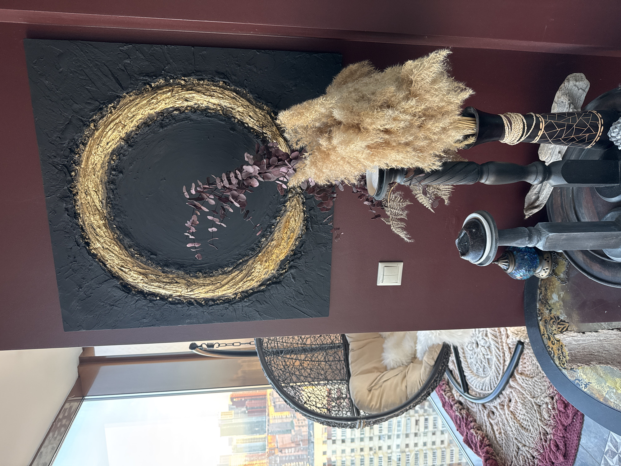 A low-angle, interior shot features a hanging chair and decorative elements within a modern setting. Outside the window is a densely populated urban landscape resembling Novosibirsk, Russia. A black-and-gold circular artwork hangs on a maroon wall. A vase of dried foliage complements the artwork. A wrought-iron, decorative table is also visible.