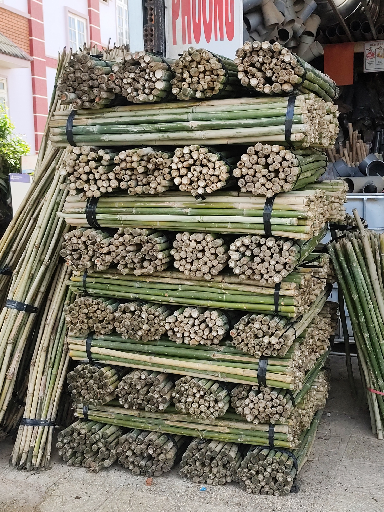 A tall stack of bundled bamboo poles is displayed on a concrete surface in Lạc Dương, Vietnam. The poles are cut into sections, bundled, and secured with black ties. The stack contains multiple tiers, each consisting of several bundles arranged horizontally. The poles exhibit a light green color, with the cut ends appearing as round cross-sections. In the background, elements of a building with a pink facade and white windows are visible, along with a sign in Vietnamese. PVC pipes and other building materials are visible in the background to the right of the stack. A smaller bundle of bamboo poles leans against the stack on the left, and another stands upright on the right.