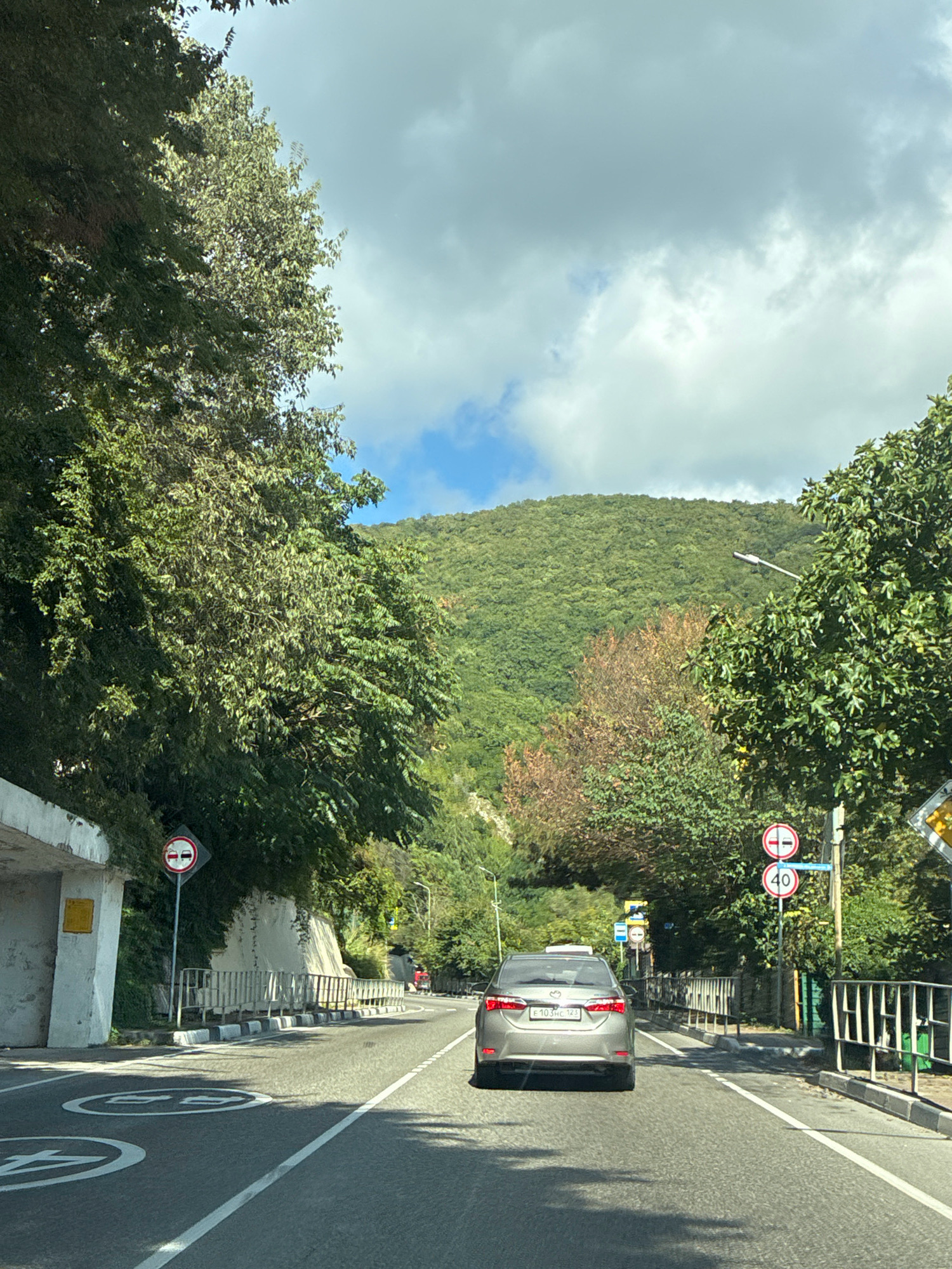 The image captures a view from a vehicle traveling on a road, likely in a mountainous or hilly area, with dense green trees lining both sides. The road curves slightly to the right. In the foreground, the road surface shows white lane markings and circular white symbols painted on it. A silver sedan is driving ahead in the same lane.
On the left side of the road, there's a concrete structure, possibly a tunnel entrance or an overhang, with a white wall and a yellow sign. Further along the left, a diamond-shaped traffic sign with a red border and two red horizontal bars within a circle is visible, indicating a restriction on passing.
On the right side of the road, there are two circular traffic signs stacked vertically. The top sign shows a red border with two red horizontal bars. The bottom sign displays a red circle with a white background and the number "40" in black, indicating a speed limit of 40. A metal fence runs along the edge of the road. Further down the road, there are more traffic lights and signs. In the distance, a red vehicle is seen.
The sky above is partly cloudy with patches of blue. The background is dominated by a lush green, tree-covered mountainside. The overall impression is a scenic drive through a vegetated area with some urban infrastructure, suggesting it could be a road leading into or out of a town or city.
Based on the provided information, this scene is located on the Lazarevskaya highway (A-147) approaching Tuapse, Russia, specifically near the entrance of the city. The mountainous terrain and coastal proximity are characteristic of this region.