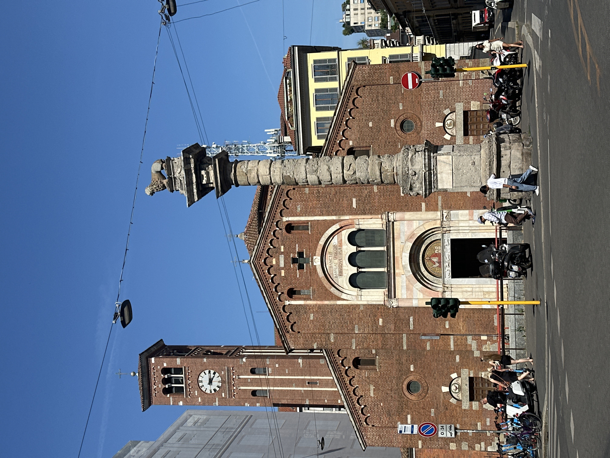 This image shows a street scene in Milan, Italy, on a bright, sunny day. A red brick church with a tall bell tower is the prominent feature. The church has arched windows and decorative brickwork. A stone column with a sculpture on top stands in front of the church. Several people are visible on the sidewalk and street, some leaning on motorcycles, others standing or walking. Motorcycles and bicycles are parked along the street. Traffic lights and street signs are also present. The sky is clear blue. The overall impression is a typical urban environment in Italy, with a mix of historical architecture and modern life.
