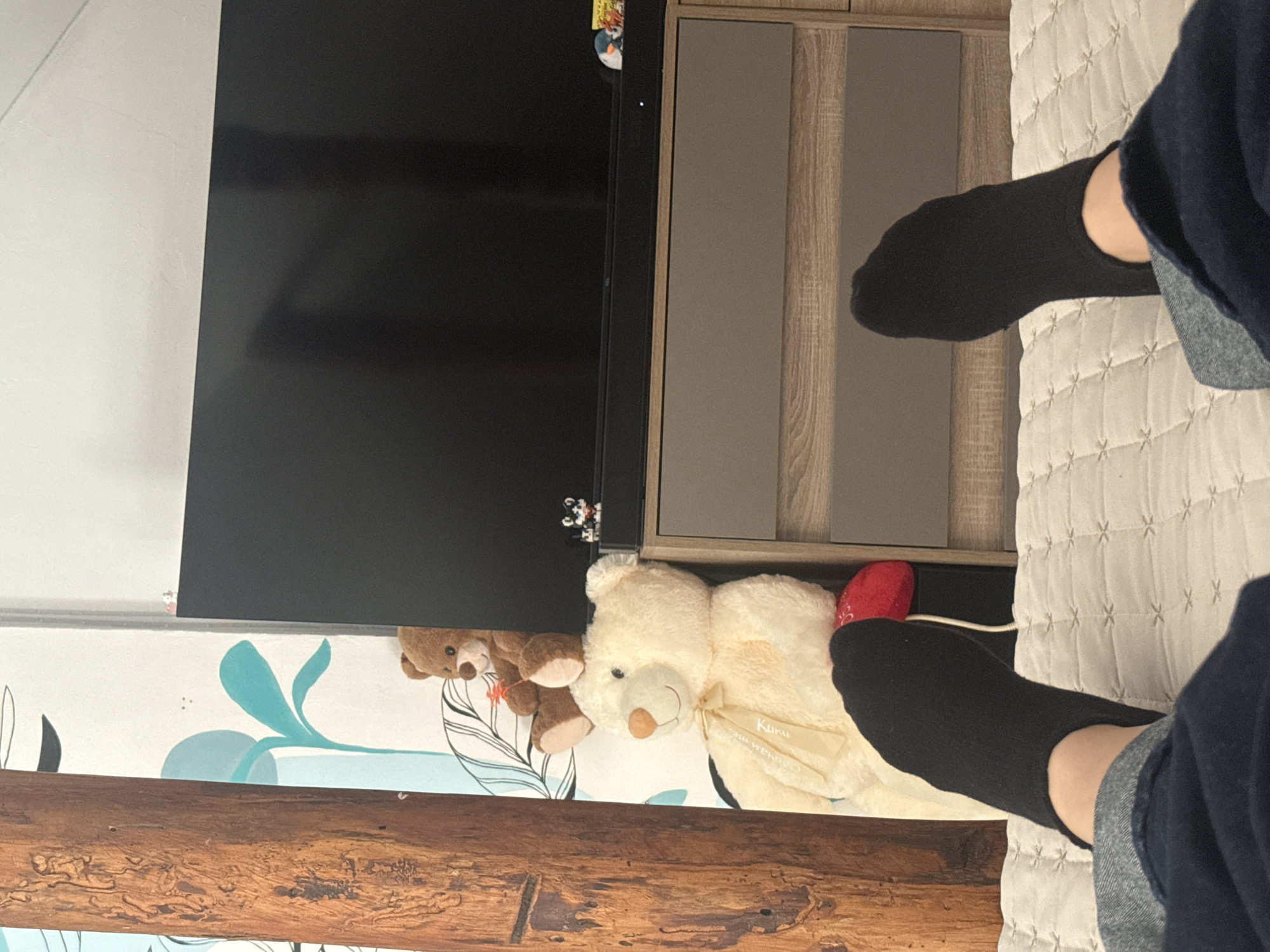 A close-up interior shot, apparently taken in Elhovo, Bulgaria, features a bed with a white quilted bedspread on the right side of the frame. A person, wearing dark-colored socks and blue jeans, is sitting or lying on the bed with their legs extended into the frame. To the left of the bed is a flat-screen television and a cabinet with wood-grain doors. A white teddy bear and a smaller brown bear are visible beneath the television. A small, red object is positioned near the white teddy bear. A wood-grained decorative element runs along the bottom left of the frame. The wall behind the furniture has a white background with a blue floral design. The shot composition suggests a casual, candid moment captured indoors.