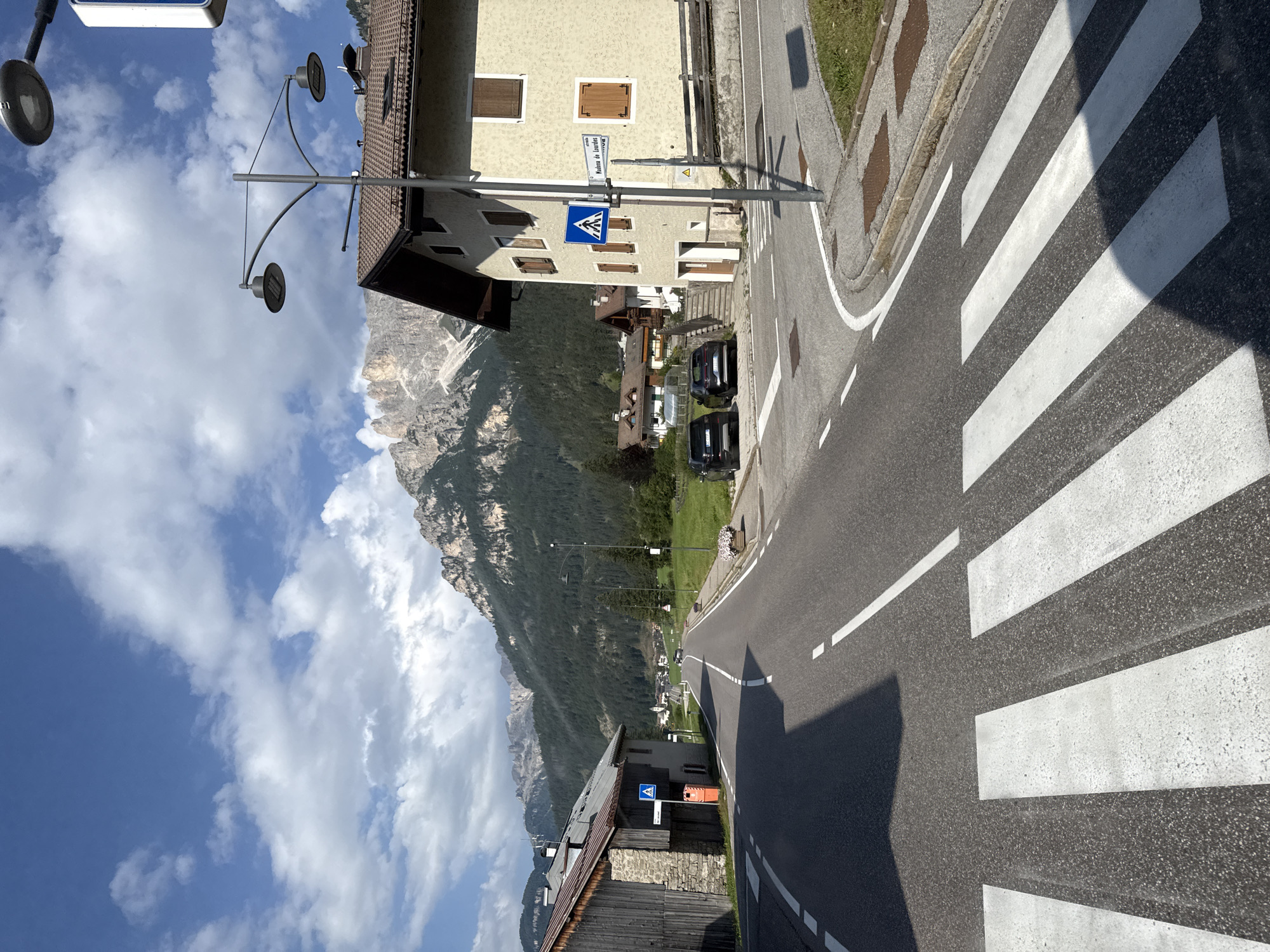 The image depicts a street scene in Mazzin - Mazin, Italy, on a sunny day with scattered clouds. A paved road with white lane markings runs diagonally from the foreground into the distance. A pedestrian crossing with white stripes is prominent in the lower portion of the frame. On the right side of the road, a light-colored building with brown shutters and a tiled roof stands. A street lamp with two lights is positioned in front of this building, along with a blue pedestrian crossing sign and a street sign that reads "Madonna de Lourdes". Two dark-colored SUVs are parked on the road in front of the building. Further down the road, more buildings and trees are visible, with a mountainous landscape covered in dark green trees dominating the background. On the left side of the road, a stone and wood building is partially visible, with a wooden fence and a blue pedestrian crossing sign. The overall environment suggests a quiet, picturesque village setting nestled in the mountains.