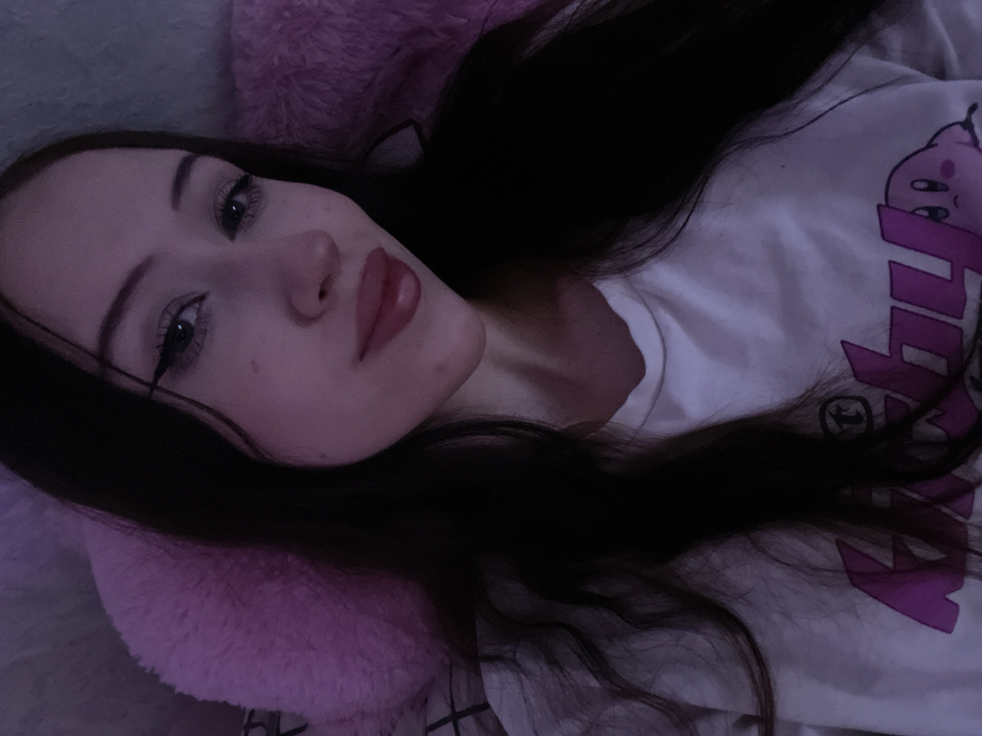 The image is a close-up selfie of a young woman with long, dark hair. She is wearing a white t-shirt with a pink cartoon character on it. The background appears to be a bed with a pink fluffy pillow. The lighting is dim, with a purple hue. There is no indication of the location being in Votkinsk, Russia.