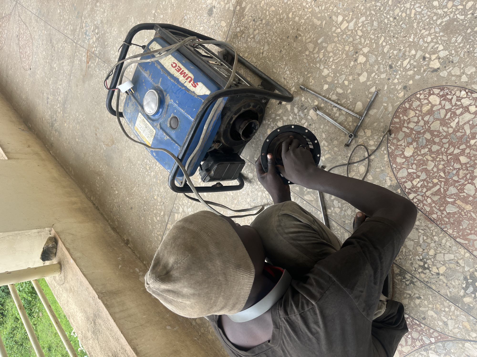 The image shows a person, likely a male, sitting on a terrazzo floor and working on a blue generator. The generator has a black metal frame and some wires are connected to it. Tools, including a wrench, are also on the floor near the generator. The person is wearing a tan cap and a dark t-shirt, and their hands are engaged in manipulating a part of the generator, possibly for repair or maintenance. The scene appears to be outdoors, with a concrete structure and some greenery visible in the background, suggesting a building's exterior or a balcony. The lighting suggests it is daytime. There is text visible on the generator, which appears to be the brand name "SUMEC". The overall impression is one of focused activity and practical work.