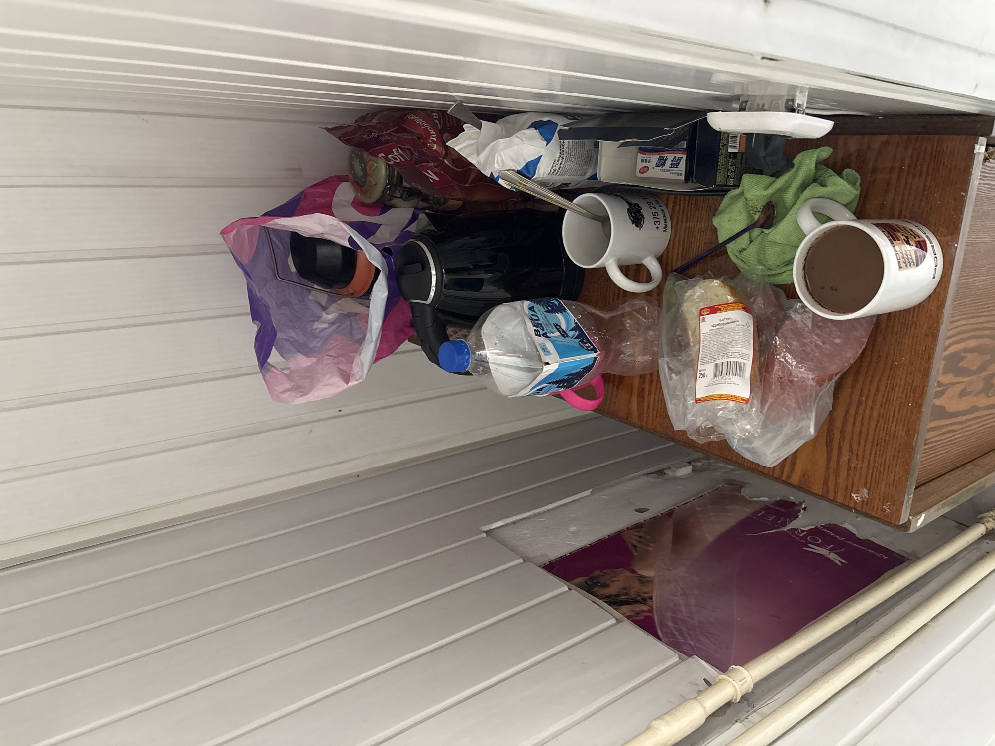 The image shows a close-up, high-angle view of a small wooden shelf against a white paneled wall. On the shelf are various items, including a black electric kettle, a bottle of "Aqua" brand water, a white mug with a logo and text that appears to be a phone number and location in Minsk, a bag of coffee, and other food items in plastic packaging. There is also a green cloth with a spoon on it and another white mug filled with a dark liquid, possibly coffee, with a Porsche logo on it. To the left of the shelf, partially visible, is a large poster with a woman's image and some text. The overall impression is that of a cluttered domestic or break area. The image does not contain any information that would indicate its location in the city of Vileyka, Belarus.