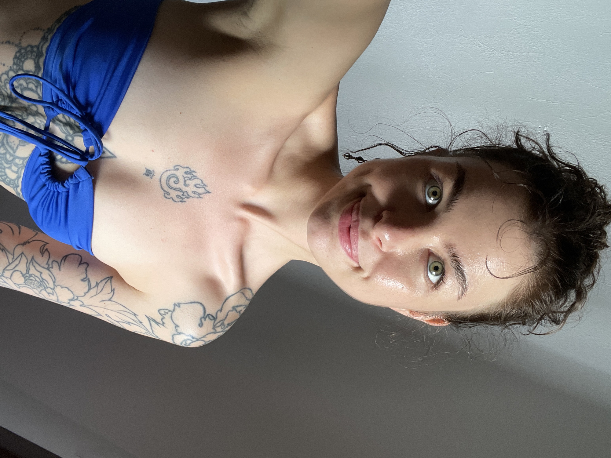 A young adult female is centered in a close-up, chest-up self-portrait. She has light skin, wet, dark, curly hair pulled back, and striking green eyes. Her face appears moist with a slight, closed-lip smile. A small, dark, dangling earring is visible on her left ear. She wears a royal blue strapless bandeau bikini top, featuring a central drawstring detail. Visible tattoos include a stylized flame design with a spiral and a Chinese character (炎) on her sternum, outline floral patterns (peonies/roses) on her right shoulder and upper arm, and intricate geometric or mandala-style line work on her abdomen below the bikini top. Her right arm is raised, suggesting she is holding the device used for the self-portrait. The background is a plain wall, light gray and textured on the left, transitioning to a smooth, dark gray shadow on the right. Lighting appears to originate from the left, illuminating the left side of her body and face while casting the right side into shadow. The image contains no visual elements to confirm or suggest the location as Boreti, Montenegro.