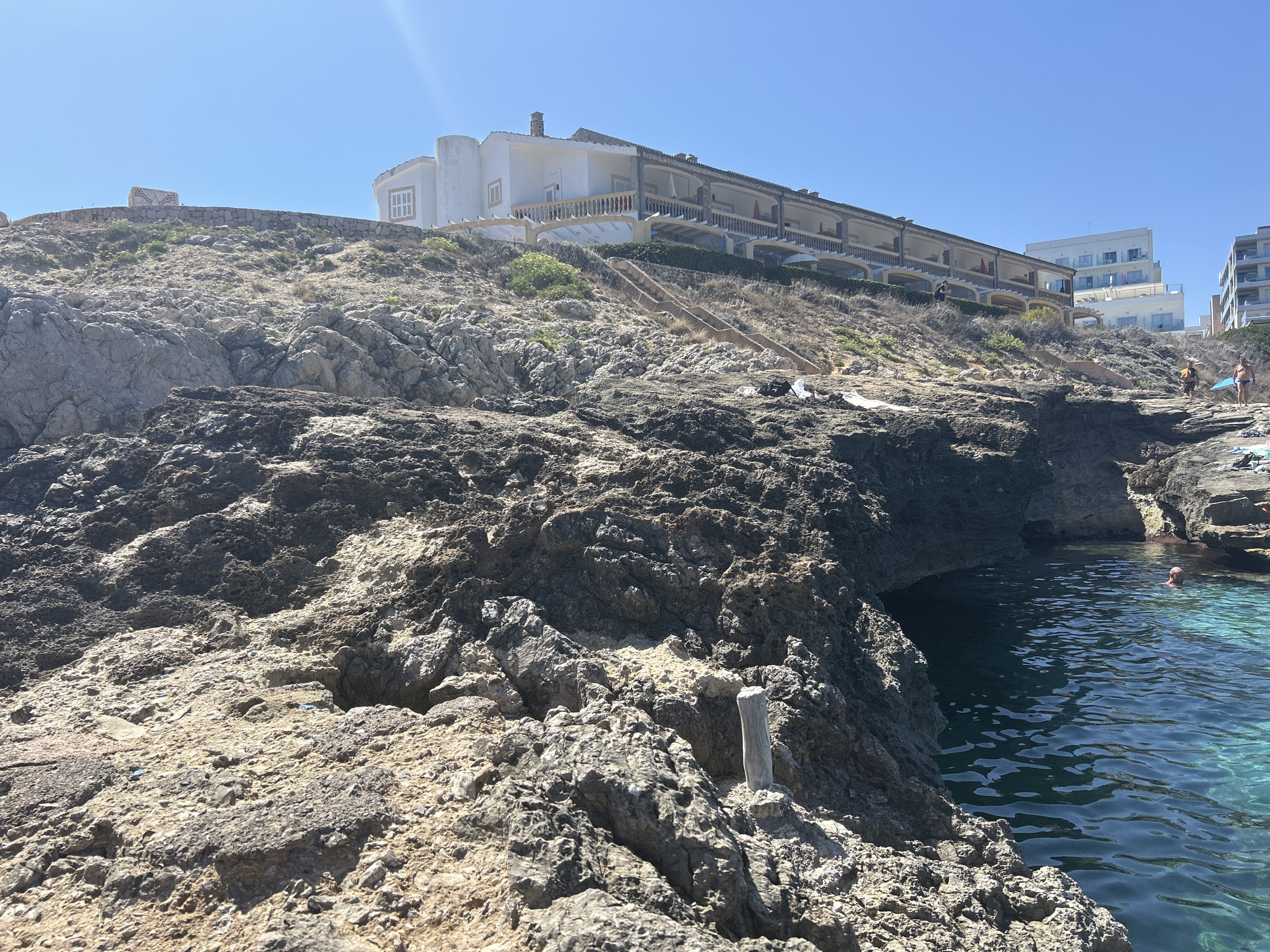 This is a photograph taken from a rocky coastline looking towards the sea and buildings situated on a hill. The foreground is dominated by rough, textured, light brown and grey rocks, with some darker, wet-looking rocks closer to the water's edge. A single, weathered wooden post stands upright among the rocks in the mid-foreground. To the right, the clear, turquoise water of the sea is visible, with a person partially submerged and a rocky overhang casting a shadow. In the background, a long, white building with multiple balconies and awnings stretches across the hillside. Further up the hill and to the right, two modern, multi-story white buildings are visible. The sky is clear and blue. The location is identified as Cala Lliteres, Spain.