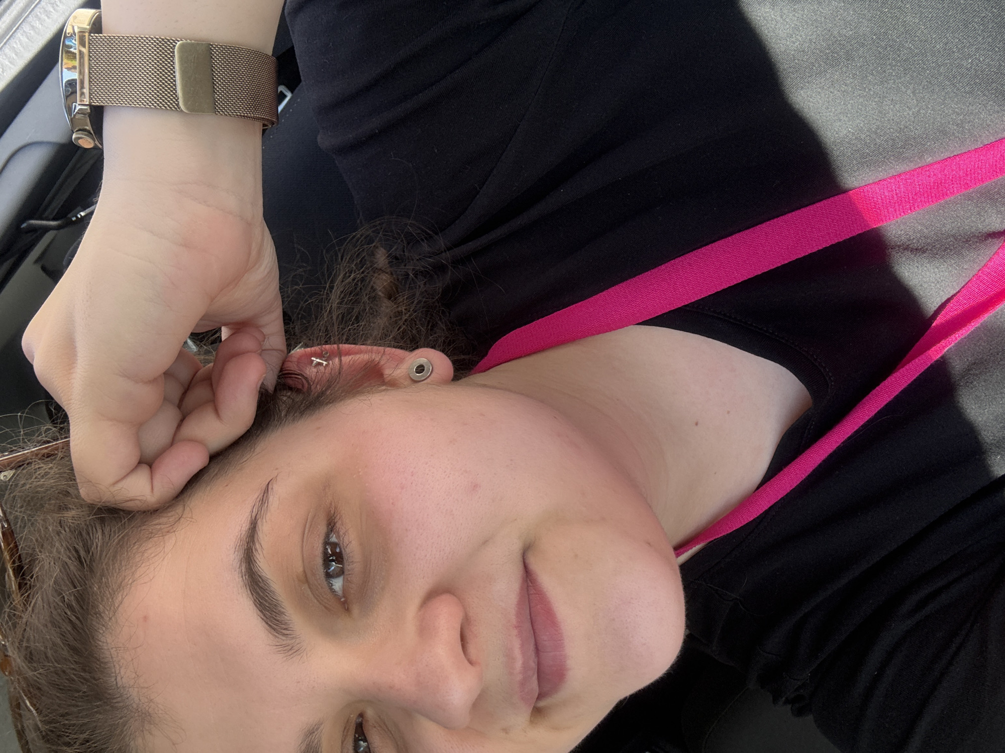 A woman is looking at the camera. She has brown hair, brown eyes, and fair skin. She is wearing a black top with a pink strap. On her left wrist, she wears a gold watch. Her left hand rests on her head. She has an earring on her ear. The location of the photo is not discernible in Plovdiv, Bulgaria.