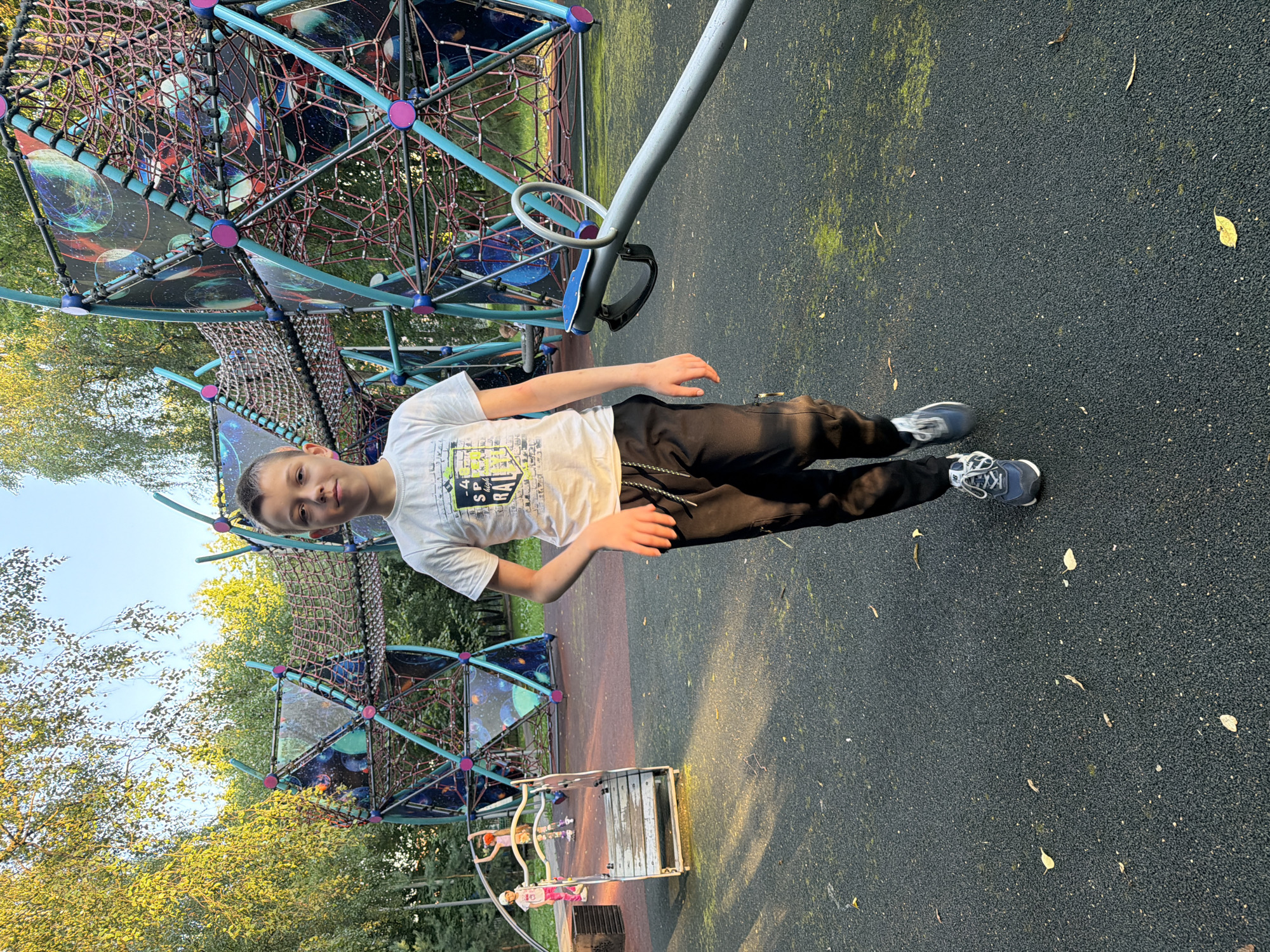 A young boy with short brown hair and fair skin is standing in a playground. He is wearing a white t-shirt with a graphic design and black pants with white stripes. He is also wearing blue and grey sneakers. The boy is facing the camera, with his left arm slightly bent and his right arm by his side.
The playground has a modern climbing structure with colorful geometric shapes and netting. A seesaw is visible in the foreground. The ground is a dark, rubberized surface with scattered leaves. In the background, trees with green and yellow foliage are visible, suggesting it is autumn. Two other children are playing on a smaller structure in the mid-ground. The lighting suggests it is late afternoon, with sunlight casting shadows on the ground.