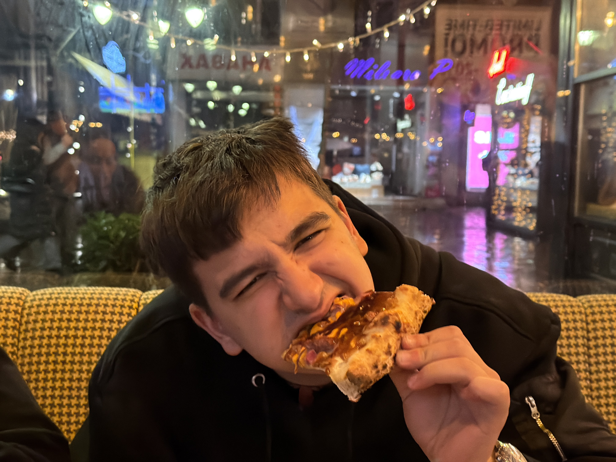 A young male, possibly in his late teens or early twenties, with short brown hair, is seated indoors at night, actively consuming a slice of pizza. He is wearing a black hooded sweatshirt and is positioned in front of a yellow and black houndstooth pattern upholstered booth or chair. The male is seen biting into the pizza slice with an exaggerated, expressive facial grimace, his eyes squinted. The pizza slice features a thick crust, a red sauce base, cheese, and what appears to be various toppings, possibly including meat and an additional sauce drizzled on top.
The setting is a restaurant or pizzeria, with a large glass window behind the male revealing an exterior street scene at night. The window reflects and transmits various city lights and neon signs. Visible reflections include string lights hanging horizontally, blurred shapes of other patrons inside the establishment, and bright, colorful neon signs from businesses across the street. These signs include portions of text, such as a purple "Wilbor" sign and a red sign with partial white text "LIMITED-TIME PROMO!" in mirror image. Other blurred lights and reflections indicate a bustling urban environment.
Based on the visible "Wilbor" neon sign and the general urban setting, the location is identified as a **Wilbor Pizza** restaurant in the city of **Sofia, Bulgaria**. Specifically, the appearance of the street and reflections align with establishments found on prominent streets like **ul. "G. S. Rakovski"**, known for its vibrant nightlife and commercial activity.