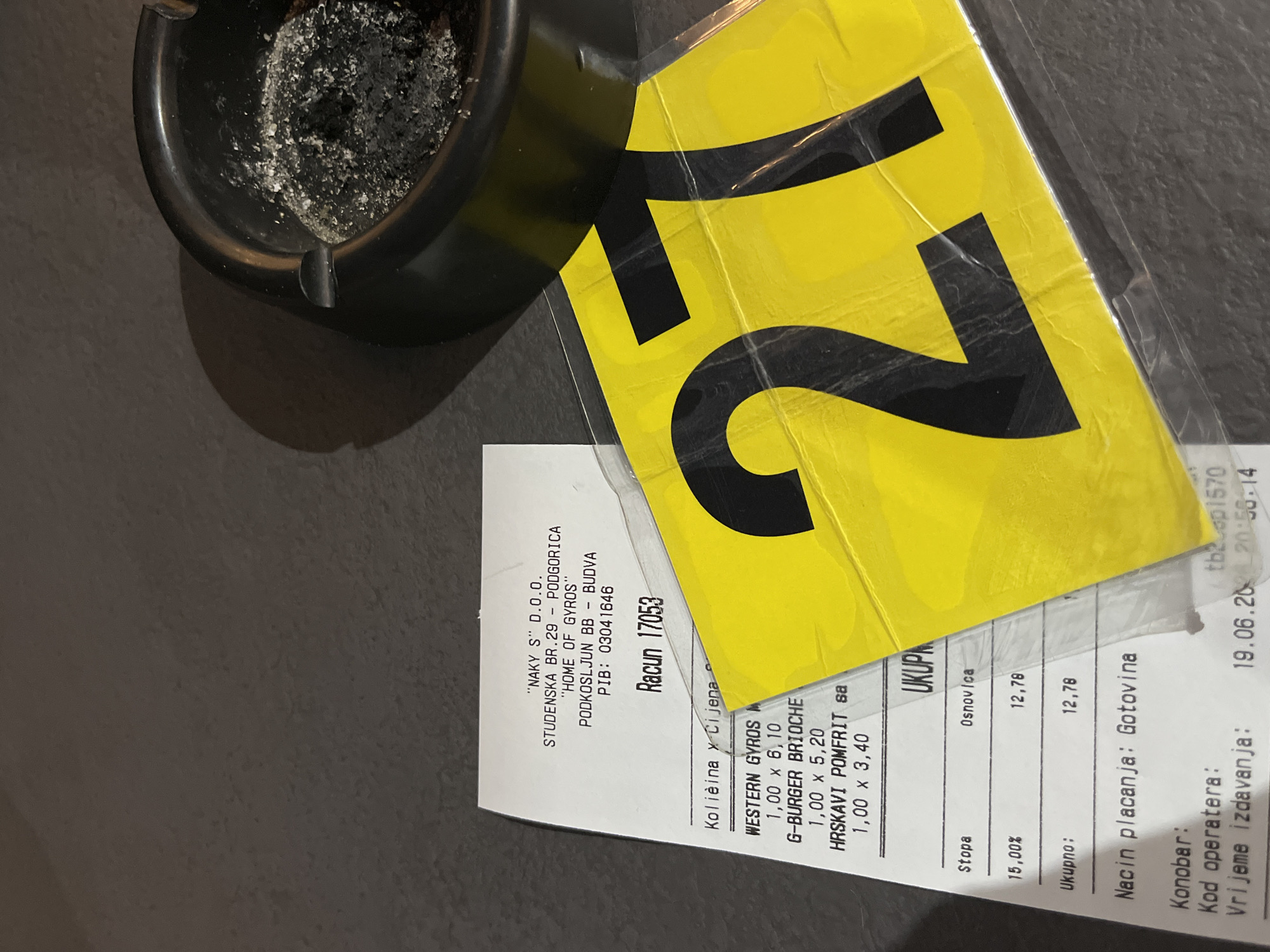 The image shows a receipt and a yellow sign with the number "27" printed on it, placed next to an ashtray filled with cigarette ash. The receipt is from "HOME OF GYROS" located at Podkosljun bb, Budva, Montenegro. The receipt details several food items including "WESTERN GYROS", "G-BURGER BRIOCHE", and "HRSKAVI POMFRIT SA", with quantities and prices listed. The total amount is 12.78. The receipt also indicates the payment method as "Gotovina" (cash) and the date and time of issue as "19.06.2023 20:56:14". The overall scene appears to be a table in a restaurant or bar.
