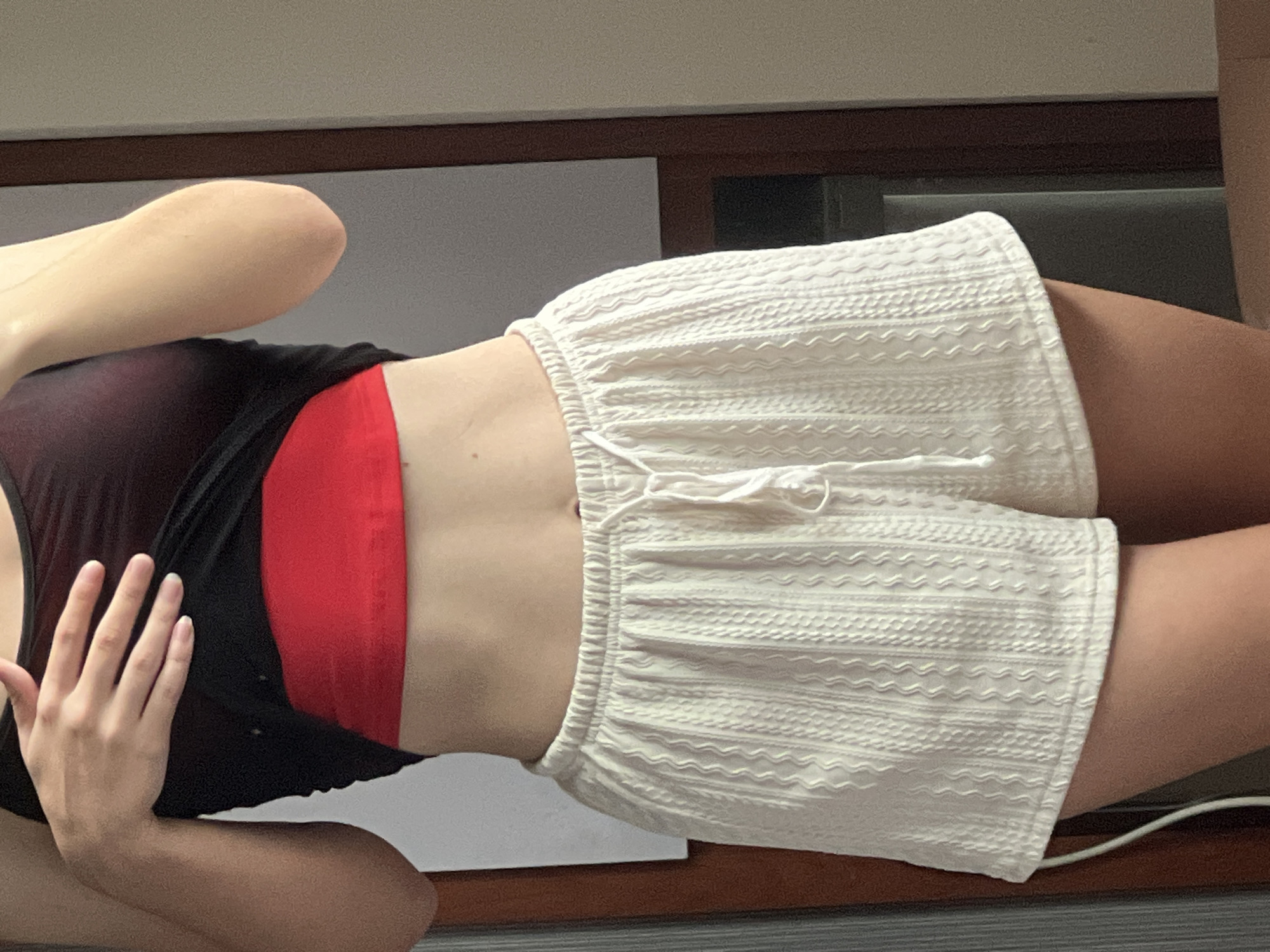 A person lies on their side wearing a black top over a red sports bra and white shorts with a drawstring waist. Their hand is partially obscuring their torso. The background consists of a dark brown surface and light gray vertical panel. The scene appears to be indoors. There is no indication of this image being in Bac Nha Trang Ward, Vietnam.