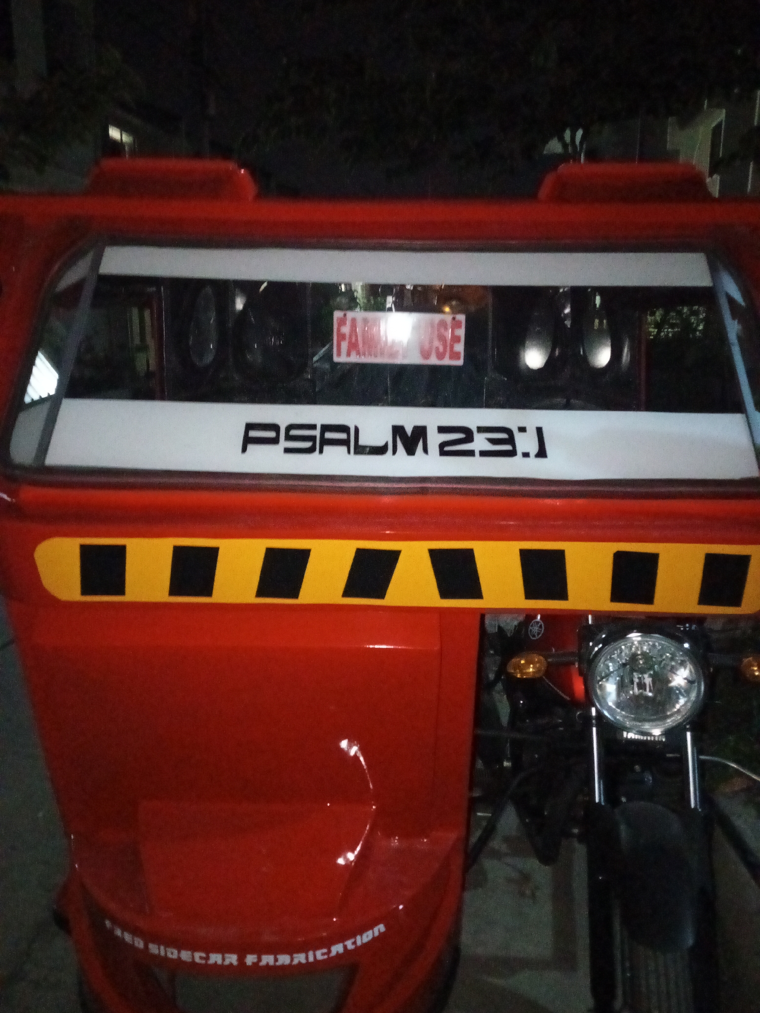 The image shows a red and black Filipino tricycle, a common mode of transportation, possibly in Davao City, Philippines. The sidecar of the tricycle displays "PSALM 23:1" in black lettering on a white background, above a yellow and black striped pattern. Text is also seen at the bottom of the sidecar reading "FRED SIDECAR FABRICATION". Behind the tricycle is darkness. The tricycle has its headlight on. A sign reads "FAMILY USE" above the scripture verse.