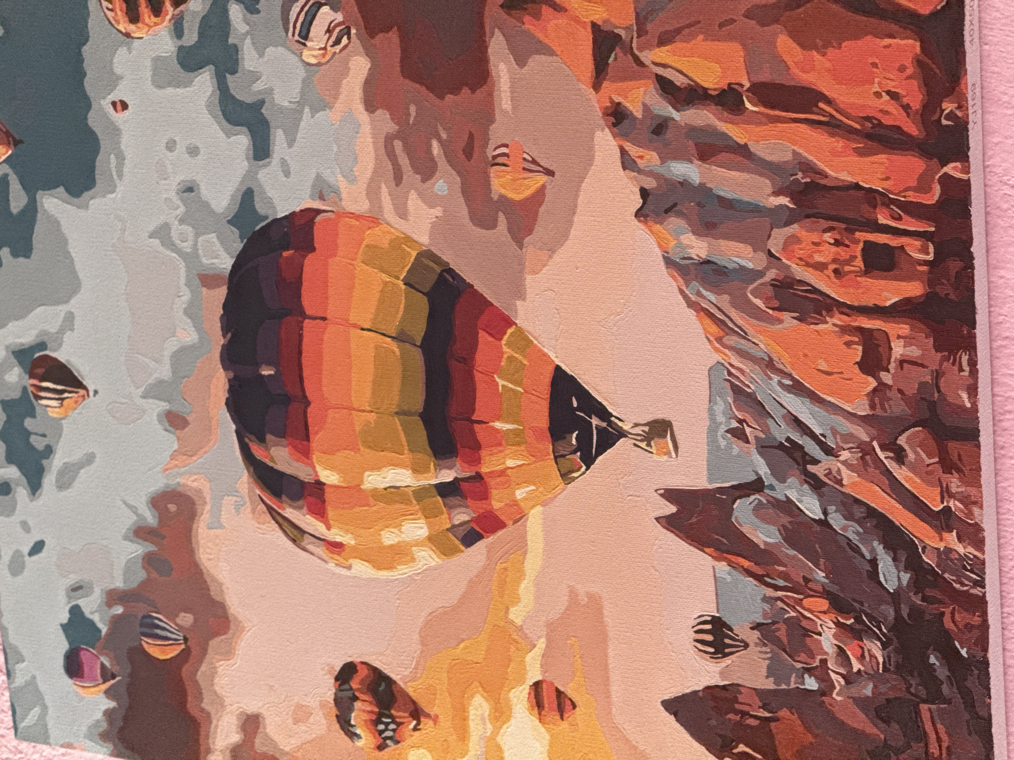 The image depicts a hot air balloon festival above a rocky landscape, likely Cappadocia, Turkey, not Basarabeasca, Moldova. The scene is dominated by a large, brightly colored hot air balloon in the foreground, with vibrant orange, yellow, and red stripes. Several other hot air balloons of varying sizes and colors are scattered across the sky, some appearing as distant specks. The sky is a mix of soft pink and grey, with hints of orange suggesting a sunrise or sunset. Below the balloons, a dramatic landscape of pointed, eroded rock formations, predominantly in shades of orange, brown, and grey, stretches across the lower portion of the image. The textures are rendered in a painted style, resembling a "paint by numbers" artwork. There are no people visible in the image. The overall impression is one of a serene and awe-inspiring aerial spectacle over a unique geological setting.