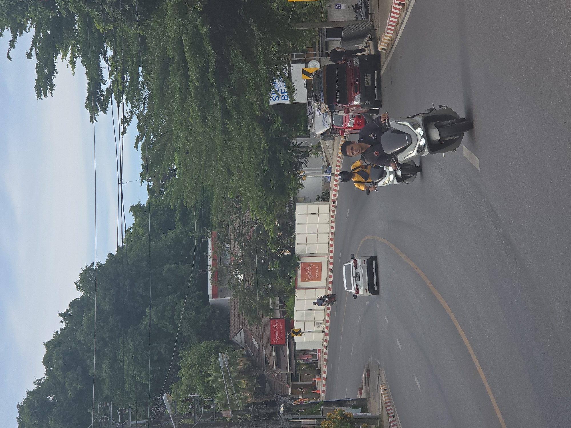 A cloudy, daytime street scene shows traffic on a paved road. On the right side of the road, a red pickup truck with a roof rack is parked, and a person in a black shirt walks past it. Further up the road, a white car is driving away from the camera, and two motorcycles are approaching. The rider of the foremost motorcycle is a man in a black shirt and dark pants. The rider of the second motorcycle is wearing a yellow shirt and a black helmet. The road is lined with trees on the left and right, and buildings are visible behind the trees. Signs for "Crystal Bay Beach Resort" are visible on the left side of the road.