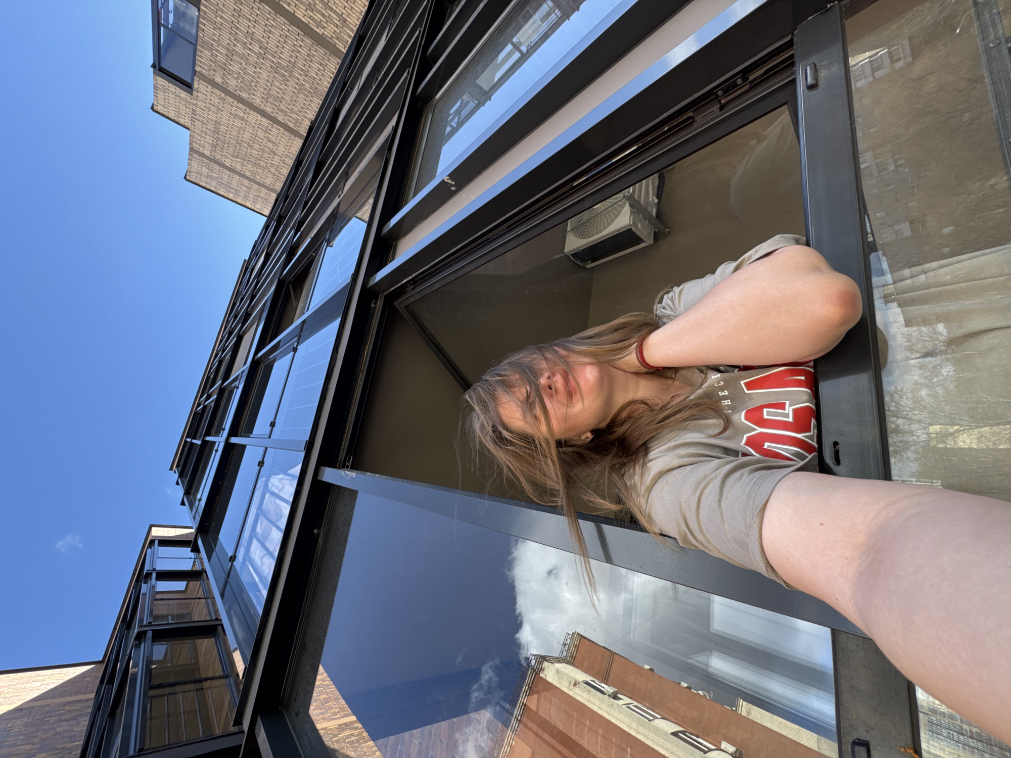 A young woman leans out of a modern building's window, captured from an upward angle against a clear blue sky. Her light brown hair partially covers her face as she looks out, resting her left arm on the dark metal window frame. She wears a light-colored t-shirt with red lettering, partially obscured.
The building exhibits a contemporary design with large glass panes and dark, angular frames, reflecting the sky and a distant, older-style reddish-brown brick building. To the left, another section of a light-colored brick building is visible against the bright sky. An air conditioning unit is seen through a window pane higher up.
The image depicts a bright, sunny day with a few scattered white clouds, suggesting midday or early afternoon. The overall atmosphere is calm and contemplative, with the woman appearing relaxed. The urban architectural elements are consistent with a city environment, such as Perm, Russia. Beyond partial lettering on the shirt, no other text is clearly legible.