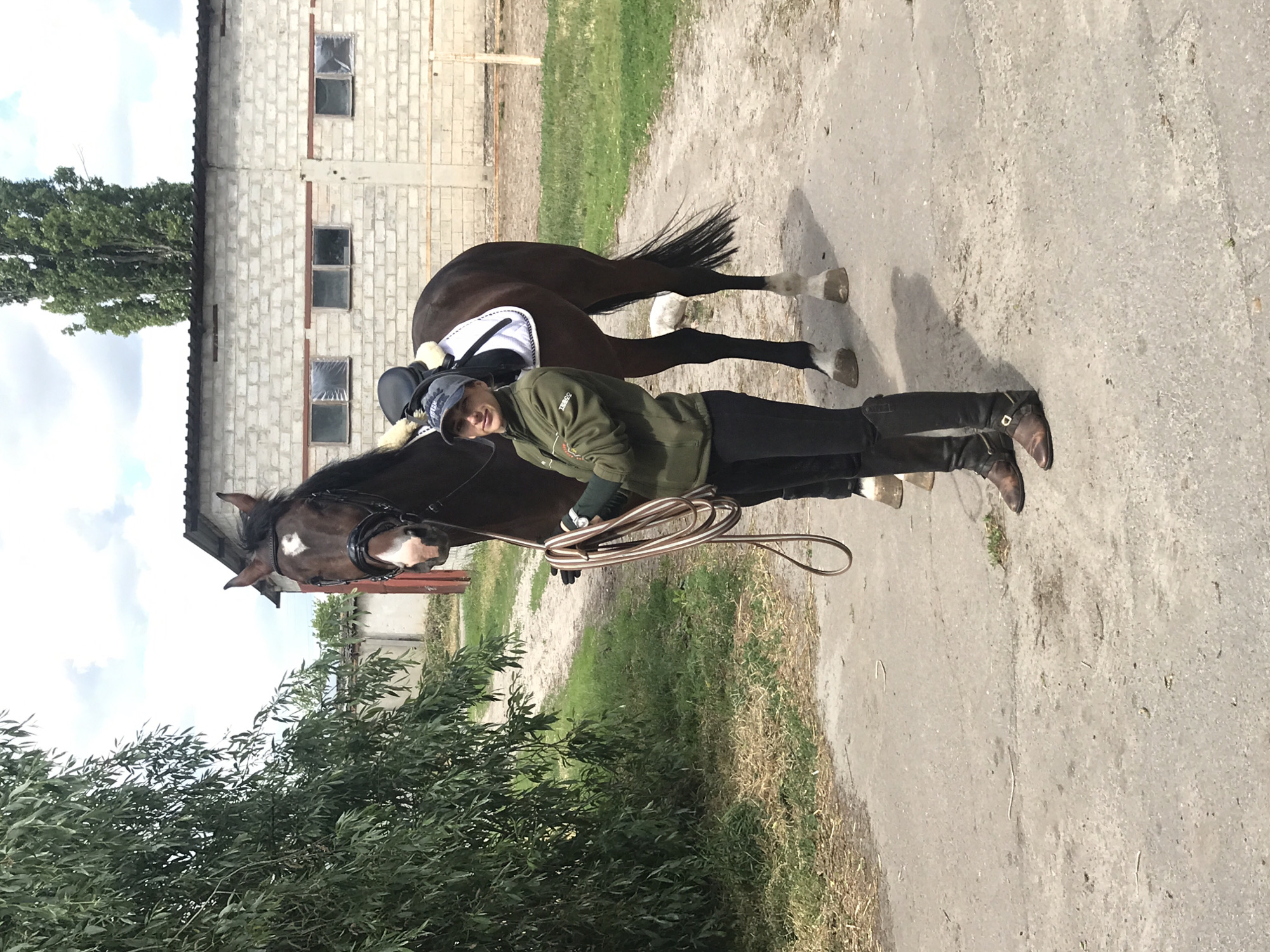 The image shows a person standing next to a horse on a paved area. The person is wearing a green jacket, dark pants, and riding boots. They are holding a lead rope. The horse is brown with a white blaze and has riding tack on. In the background, there is a building with a light-colored brick facade and green grass. There is also some vegetation in the foreground. The location is an outdoor setting, likely a stable or rural area, in the city of Unknown, Unknown.