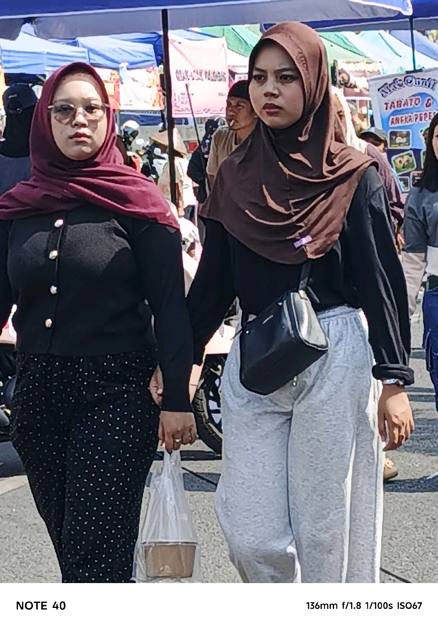 Two women are walking side-by-side in what appears to be an outdoor market in Desa Tegalreja Kecamatan Cilacap Selatan, Indonesia. The woman on the left is wearing a dark, long-sleeved shirt with gold buttons over a black skirt with white polka dots. She has on glasses and a maroon hijab. She is carrying a clear plastic bag with a brown container inside. The woman on the right is wearing a black long-sleeved shirt and light gray sweatpants. She has on a brown hijab and a black crossbody bag. Both women are looking forward, and appear to be walking away from the camera. In the background, there are market stalls with various goods and people. The ground is paved with asphalt. The image is captured with a NOTE 40 camera at 136mm, f/1.8, 1/100s, and ISO 67.