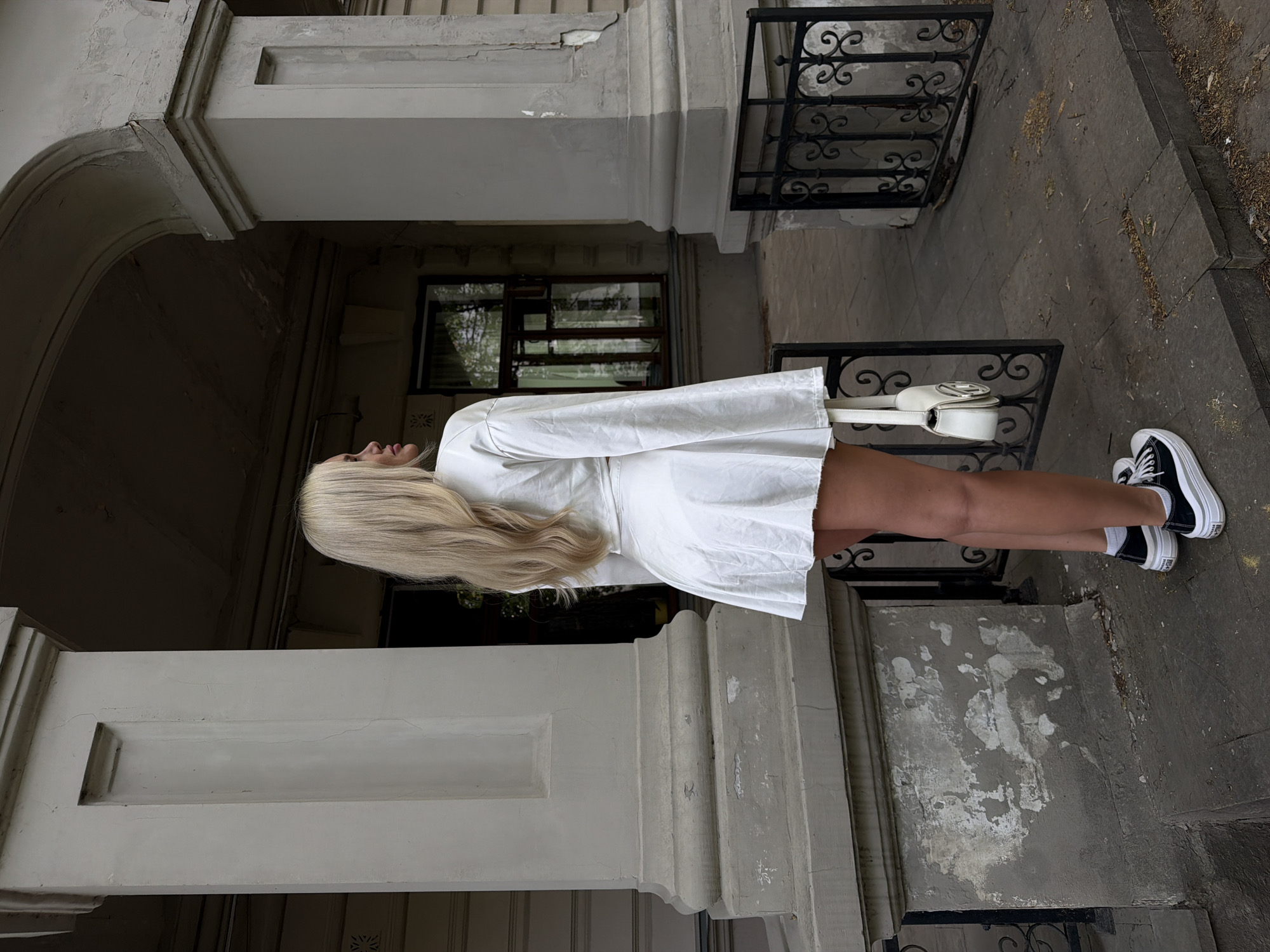 A young, light-skinned woman with long blonde hair is standing on the doorstep of a building in Kazan, Russia. She is wearing a short white dress and black and white platform sneakers with white socks. She is carrying a small white purse. The building has ornate architectural details, including railings, pillars, and a window. The doorstep and the surrounding area are made of stone.