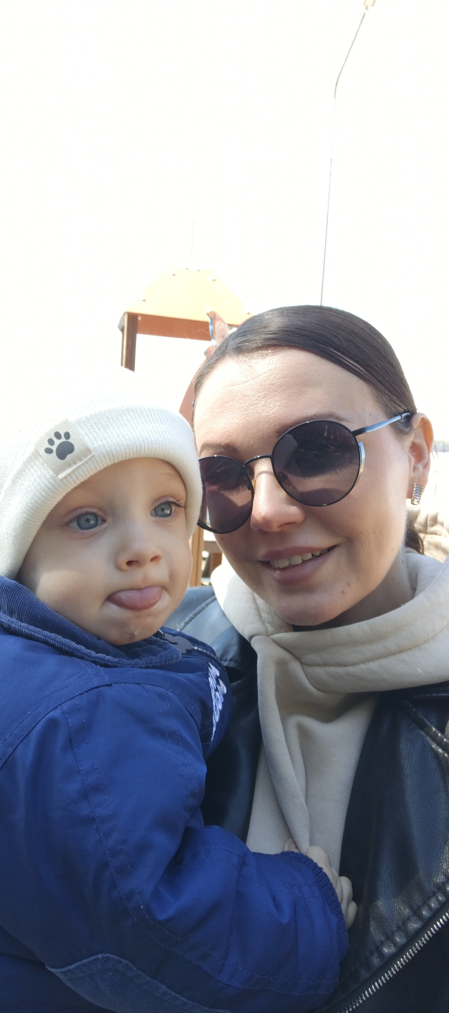 A woman and a young child are pictured in a close-up, outdoor selfie. The woman wears dark-framed sunglasses, a light-colored scarf, and a black leather jacket. The child wears a blue jacket and a light-colored knit hat with a paw print on the brim and is sticking their tongue out slightly. The background appears to be a playground area, with a partially visible structure. The location is identified as Chaikovsky, Russia.