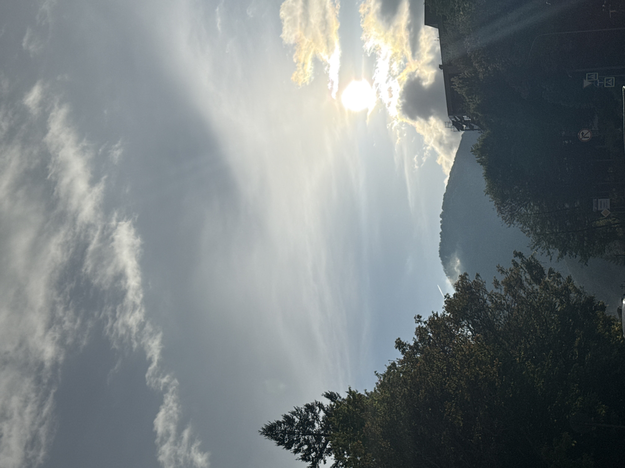 The image shows a view of the sky with clouds and the sun in Krasnaya Polyana, Russia. The sun is partially obscured by clouds. Below the sky, there is a forested mountain slope. There are trees in the foreground. There are road signs visible on the right.