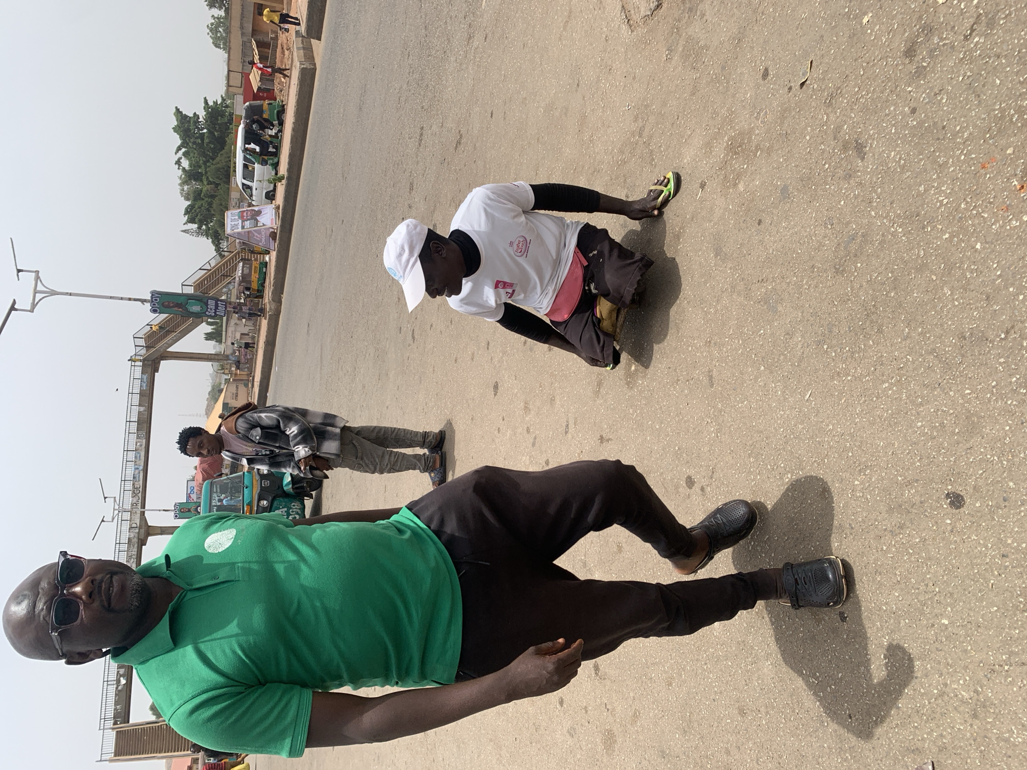 The image depicts a street scene in Jos, Nigeria. In the foreground, a dark-skinned man in a green polo shirt and black pants lies on the paved road, facing the camera, with dark sunglasses on his face. Behind him and to the right, another man wearing a white T-shirt, black shorts, and a white baseball cap kneels on the ground, facing right. Behind them, another man wearing a grey jacket and black and white checkered flannel stands on the sidewalk, looking on. A footbridge is in the background, along with several cars and people further off in the distance.