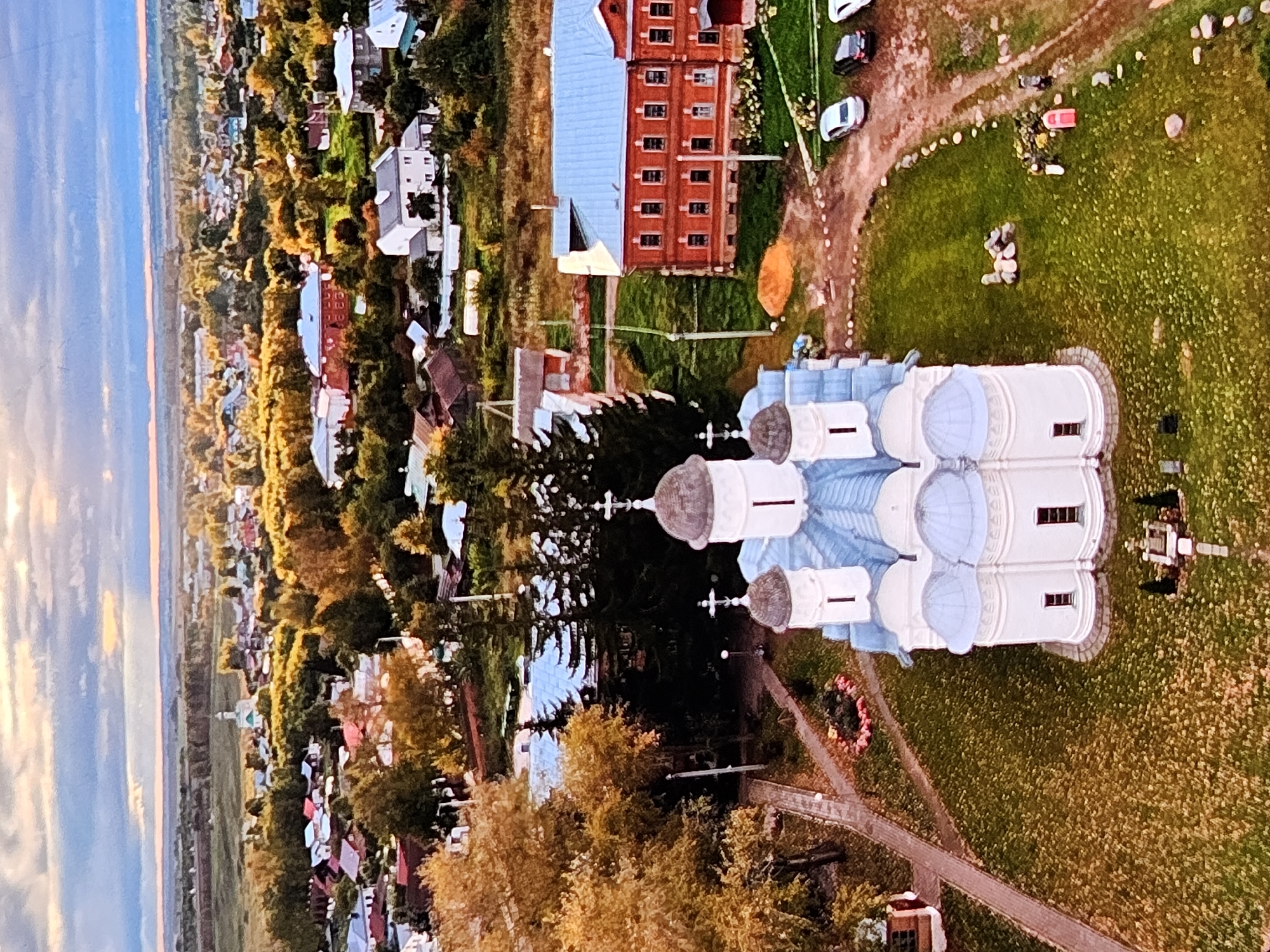 This is an aerial drone photograph of the Transfiguration Cathedral in the city of Vladimir, Russia, taken at golden hour. The cathedral, a white stone structure with light blue domes, is prominently featured in the foreground. Surrounding the cathedral is a lush green lawn, and pathways lead to its entrance. Adjacent to the cathedral is a large, red brick building with a light blue roof. The background reveals a sprawling cityscape characterized by a mix of residential houses with colorful roofs, trees displaying autumn foliage in shades of yellow and orange, and distant fields stretching to the horizon under a cloudy sky with hints of sunset colors. Several cars are parked near the red brick building, and a few individuals are visible walking on a dirt path. The overall scene depicts a peaceful, semi-rural setting within a historic Russian city.