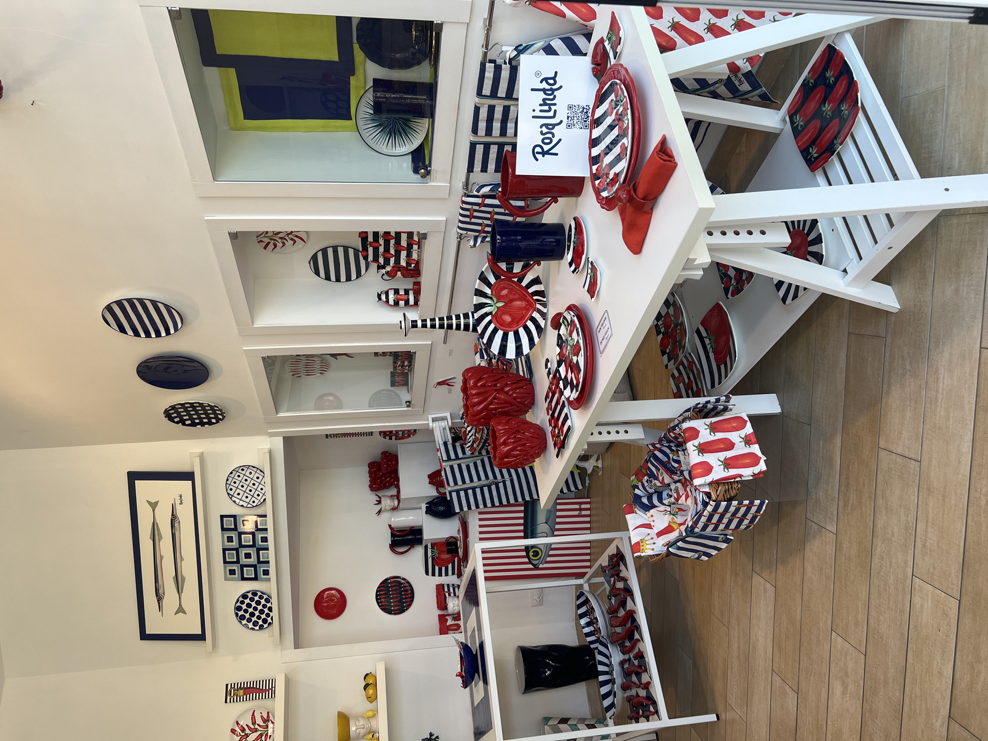 The image depicts the interior of a retail establishment, likely a shop specializing in tableware and home decor, located in the city of Sorrento, Italy. The store is brightly lit, with white walls and a wooden floor. Several display shelves and tables are arranged throughout the space, showcasing a variety of items.
The merchandise is predominantly red, white, and blue, with many pieces featuring nautical or food-related motifs, such as fish, tomatoes, and stripes. Notable items include ceramic plates, bowls, pitchers, vases, and decorative objects. A prominent display on the right features a sign with the text "Rosa Linda" and a QR code.
On the left side of the image, a wall display includes framed artwork of two fish, along with several decorative plates. A white shelving unit below holds more plates and a tall black vase.
In the center and right of the image, two white tables are set up with more tableware. The front table has various plates, napkins, and pitchers. A basket filled with folded cloths is placed on the floor in front of one of the tables.
The overall impression is one of a well-organized and visually appealing retail environment, emphasizing a specific aesthetic. There are no people visible in the image.