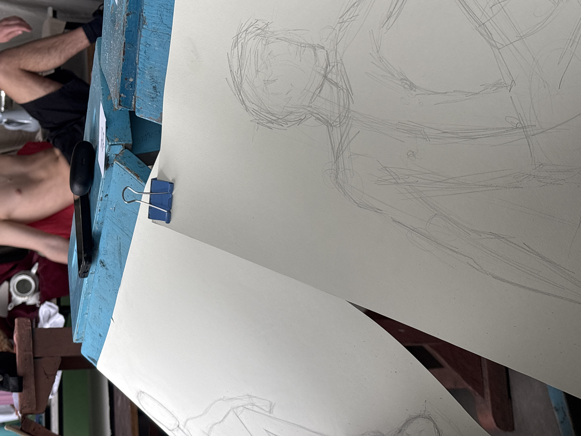 A photograph shows a still life arrangement likely depicting an art class or drawing session, likely located in Troyan, Bulgaria. Two sheets of paper are clipped to a blue painted wooden surface, each containing rough pencil sketches of human figures. Parts of a person are visible in the background to the left, torso and legs dressed in black shorts. Adjacent to the paper is a black electronic device and a binder clip secures the pages in place. In the background other objects such as a can and rags and part of a wooden stool are seen. The image provides a tilted, close-up perspective of the drawing setup.