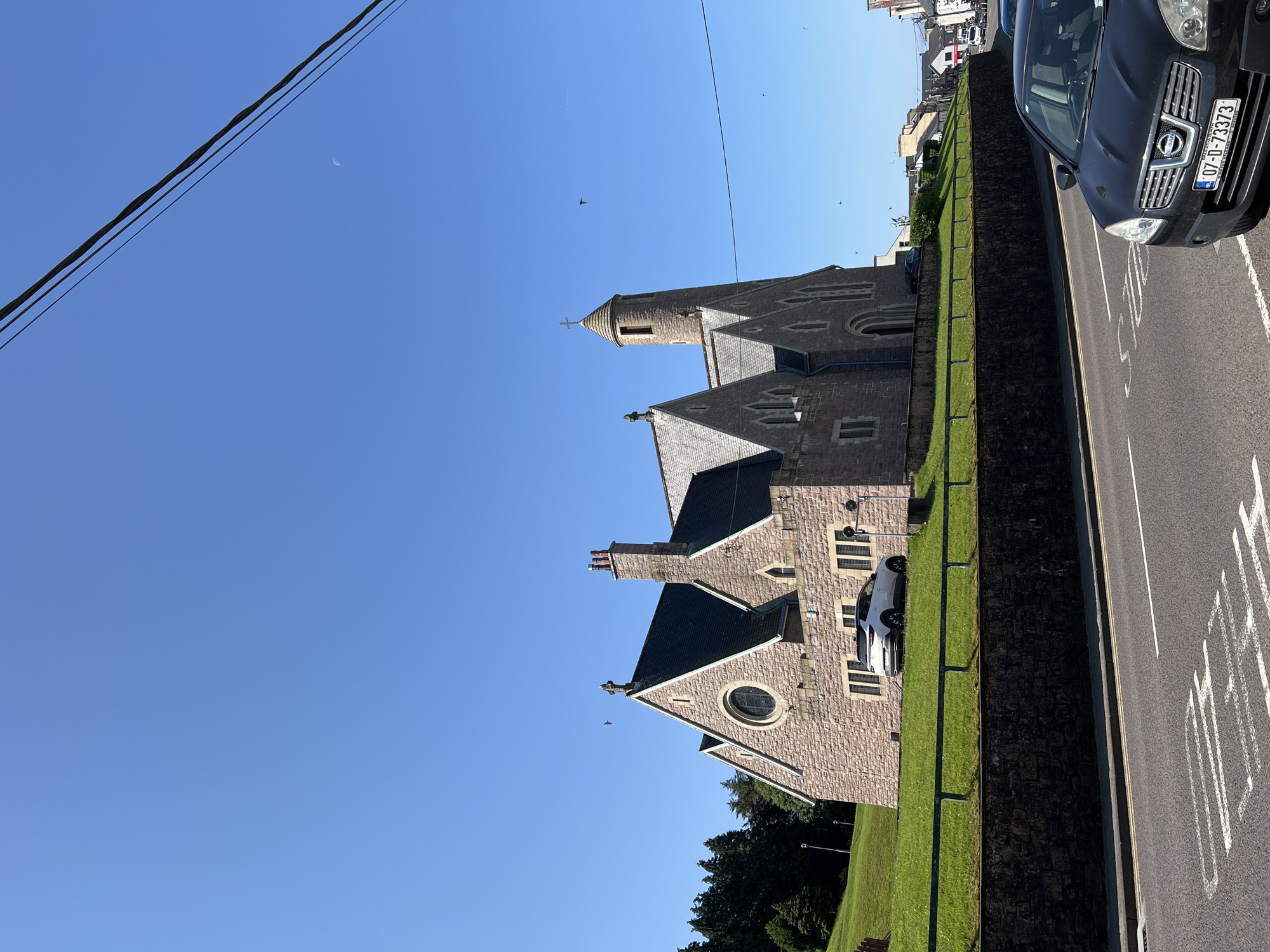 This image shows a street view in the city of Donegal, Ireland, on a clear, sunny day. In the foreground, a black Nissan Qashqai is parked on the right side of the road. The road has white lane markings and a pedestrian crossing marked with white lines.
Dominating the background is a stone church with a pitched roof and a circular window on its facade. The church has a tall, slender tower with a conical roof on its right side. A white car is parked on the grass slope in front of the church. The slope is bordered by a low, dark stone wall. Beyond the church, a line of buildings and trees can be seen. The sky is a clear blue with a few birds visible in flight and a crescent moon. Overhead, power lines are visible stretching across the frame.