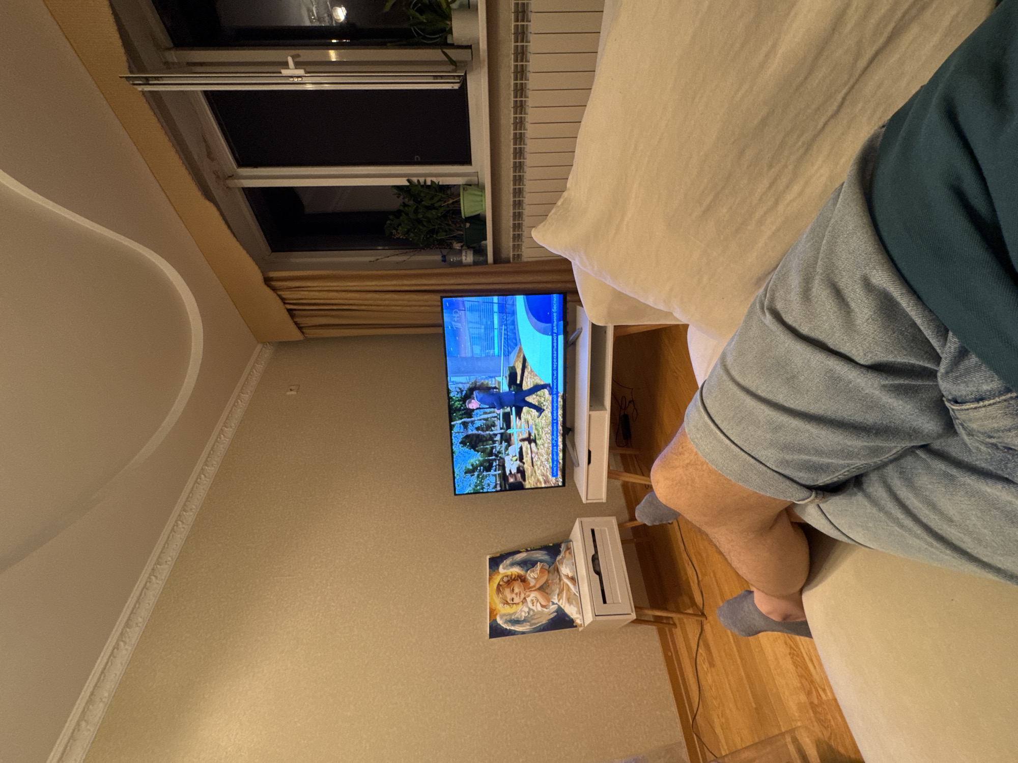 The image depicts an indoor scene, likely a living room, viewed from the perspective of someone reclining on a couch. In the foreground, the legs of a person wearing grey socks and denim shorts are visible, crossed. To the left, a small white side table with wooden legs holds a framed painting of an angel. In front of the table, a dark electrical cord runs across the wooden floor. To the right, a flat-screen television displays an image of a man in a suit walking outdoors, with text overlaid in a foreign language, suggesting a news broadcast or similar program. The television is placed on a similar white stand. Behind the television and table, a window with beige curtains is visible. The window is open, revealing the dark night sky outside. A radiator is present below the window. Several potted plants are placed on the windowsill. The walls are covered in textured wallpaper, and a decorative cornice runs along the top of the wall and ceiling. A curved line is visible on the ceiling, suggesting a decorative element or a type of ceiling construction. The overall impression is one of a relaxed evening at home. There is no specific visual information in the image that would allow for the identification of the city of Almaty, Kazakhstan.