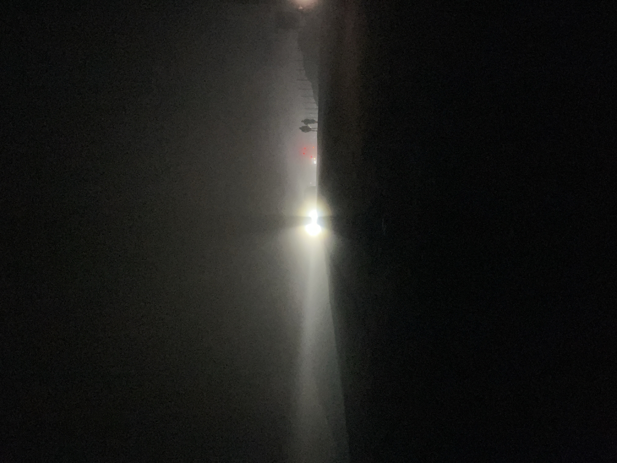 The image depicts a foggy, dark outdoor scene. In the distance, a bright light source, likely headlights from a vehicle, illuminates the mist, creating a hazy glow. Two figures, silhouetted and indistinct, stand near the light source. Further in the background, faint red lights are visible, possibly from another vehicle. The terrain appears to be a gentle slope, covered in darkness. The overall impression is one of low visibility due to heavy fog. No specific details are visible to suggest the location in Gomismta, Georgia.