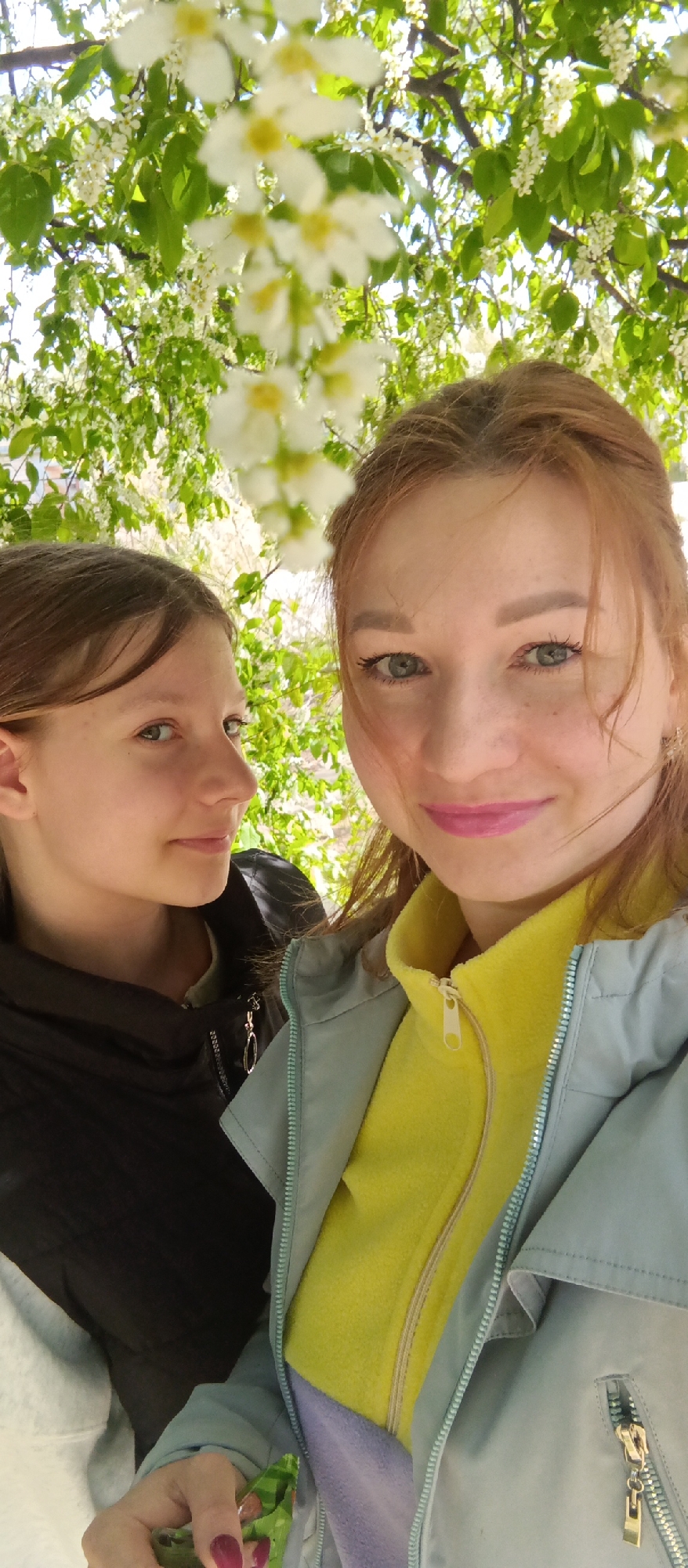 Two females are visible in the image. One female, positioned on the left, has brown hair and is wearing a black jacket. The second female, located on the right, has red hair and is wearing a yellow fleece jacket underneath a light blue jacket. Both females are looking at the camera. The background consists of white flowers and green leaves, possibly from a tree. It is daytime. The location is unknown.