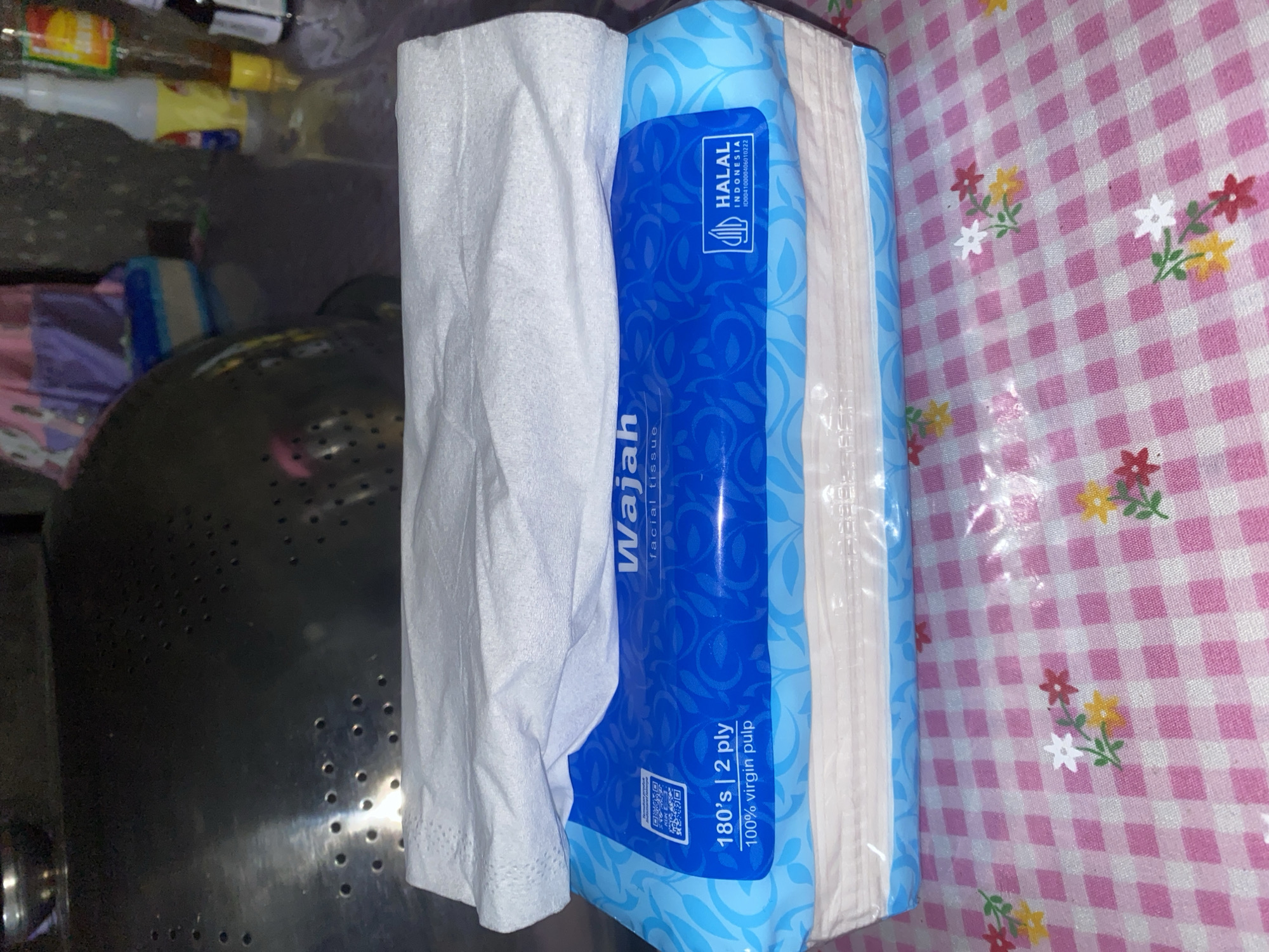 The image shows a stack of "Wajah" brand facial tissues on a pink and white checkered surface, likely a tablecloth. The tissue packaging is blue with white swirling patterns and features text indicating "180's | 2 ply" and "100% virgin pulp." A "HALAL INDONESIA" certification is also visible on the packaging. A white tissue is draped over the top of the stack. In the background, out of focus, is a metal colander or strainer with perforations. To the right of the colander, other objects are visible, including bottles and a portion of what appears to be clothing or fabric. The environment appears to be indoors, possibly a kitchen or dining area. The image does not contain any explicit information about the city of Bontoharu, Indonesia, or any people present.