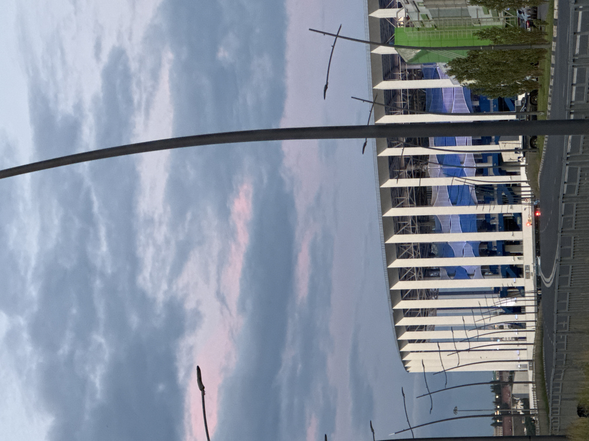The image shows a view of the Nizhny Novgorod Stadium, also known as the Nizhny Novgorod Arena, located in Nizhny Novgorod, Russia. The stadium is a large, modern structure with a distinctive white facade composed of vertical supports. The sky above is cloudy, with hints of pink and purple, suggesting either dawn or dusk. Several tall lampposts are visible in the foreground and midground, some curving dramatically. A road runs in front of the stadium, with a fence separating it from a grassy embankment. A few vehicles are present on the road and parked near the stadium. The stadium itself appears to be under construction or undergoing renovations, as indicated by the netting draped over some sections and a large green tarpaulin on the right side. The overall impression is one of an urban scene at twilight, with a prominent sporting venue as the central subject.