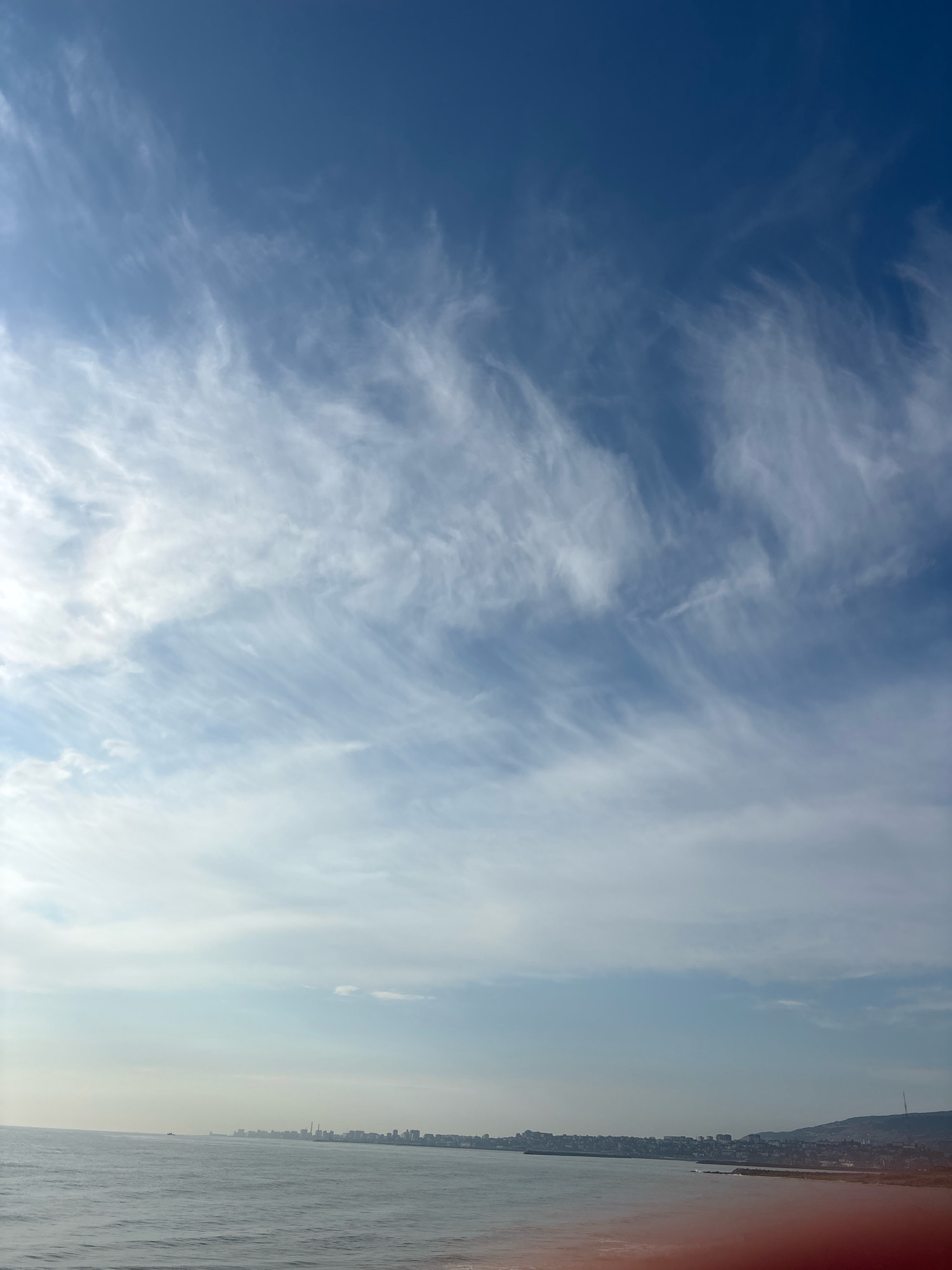 The image shows a panoramic view of the Caspian Sea coastline, likely in Makhachkala, Russia. The sky is a clear, deep blue, with wispy cirrus clouds stretching across the upper half of the frame. The sun is bright, casting a strong light on the scene.
In the distance, a city skyline is visible along the coast, characterized by a mix of low-rise buildings and taller structures, including what appear to be industrial chimneys and a prominent radio tower on a hill. The sea is calm, with gentle waves lapping at the shore. The water has a grayish-blue hue, reflecting the sky. The shoreline is a mix of rocky outcrops and what seems to be a built-up area. In the lower right corner of the image, a red-tinted glare suggests a lens flare or reflection. There are no people or immediate actions depicted in the image.