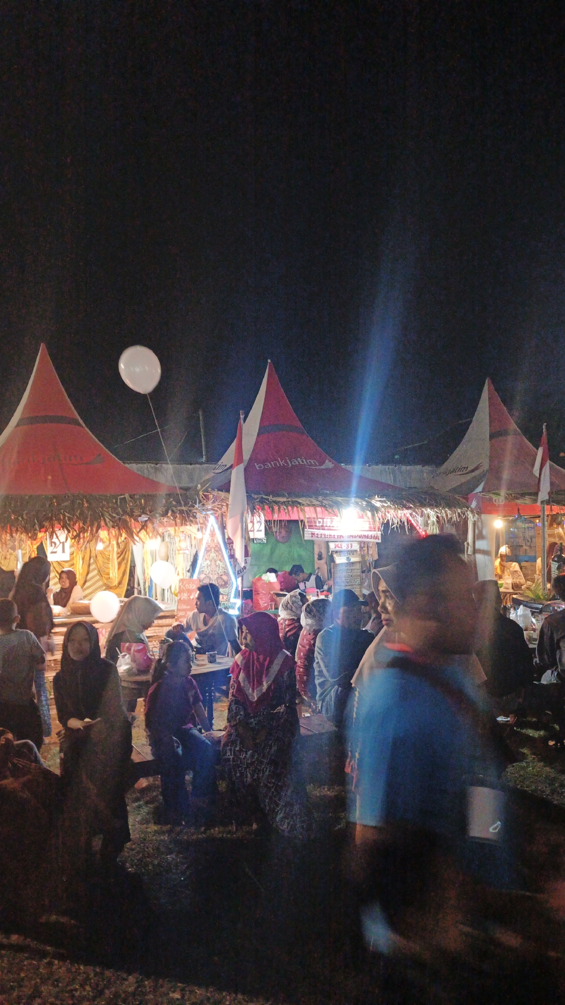 The image depicts an outdoor night market scene in Ketemasdungus, Indonesia. Several red and white striped tents, possibly stalls, are set up, some with the "bankjatim" logo visible. White balloons are suspended in the air. People are gathered around the stalls, some appearing to be customers and others vendors. The lighting is artificial, creating a festive atmosphere. The ground appears to be covered in grass and dirt. The overall impression is one of a lively community event.