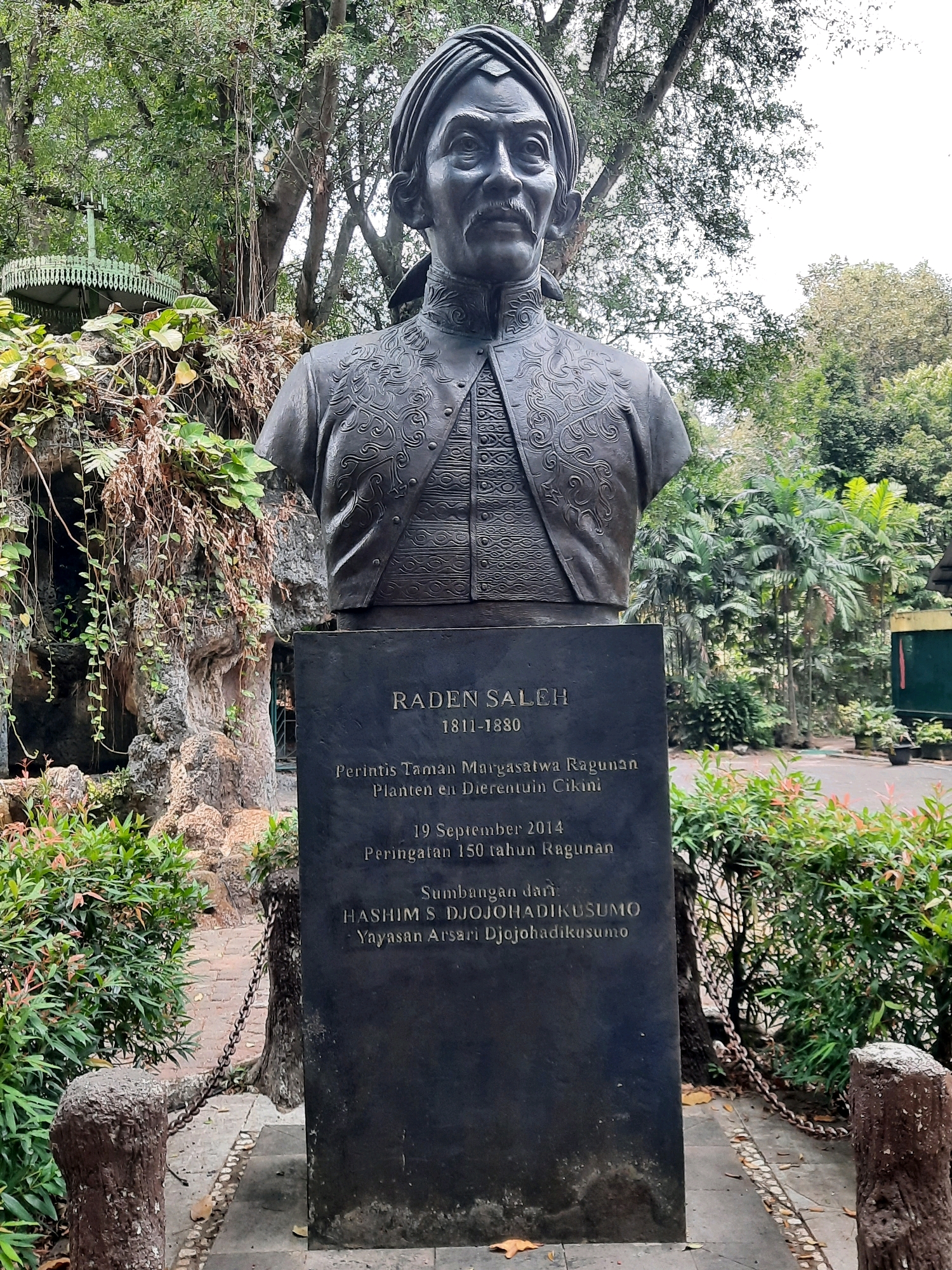 A bronze bust of Raden Saleh, a prominent Indonesian painter, stands on a dark stone pedestal in Ragunan Wildlife Park, South Jakarta, Indonesia. The pedestal features an inscription detailing Raden Saleh's life dates (1811-1880), his role as a pioneer of Ragunan Wildlife Park, and a commemoration of the park's 150th anniversary in September 2014. The inscription also credits the contributions of Hashim Djojohadikusumo and Yayasan Arsari Djojohadikusumo. The bust depicts Raden Saleh with a turban and traditional attire. The surrounding environment includes lush green trees, foliage, and a paved walkway with stone posts and chains. In the background, a vintage green structure, possibly an old gazebo or shelter, is visible amidst the trees. The overall scene conveys a sense of historical tribute within a natural setting.