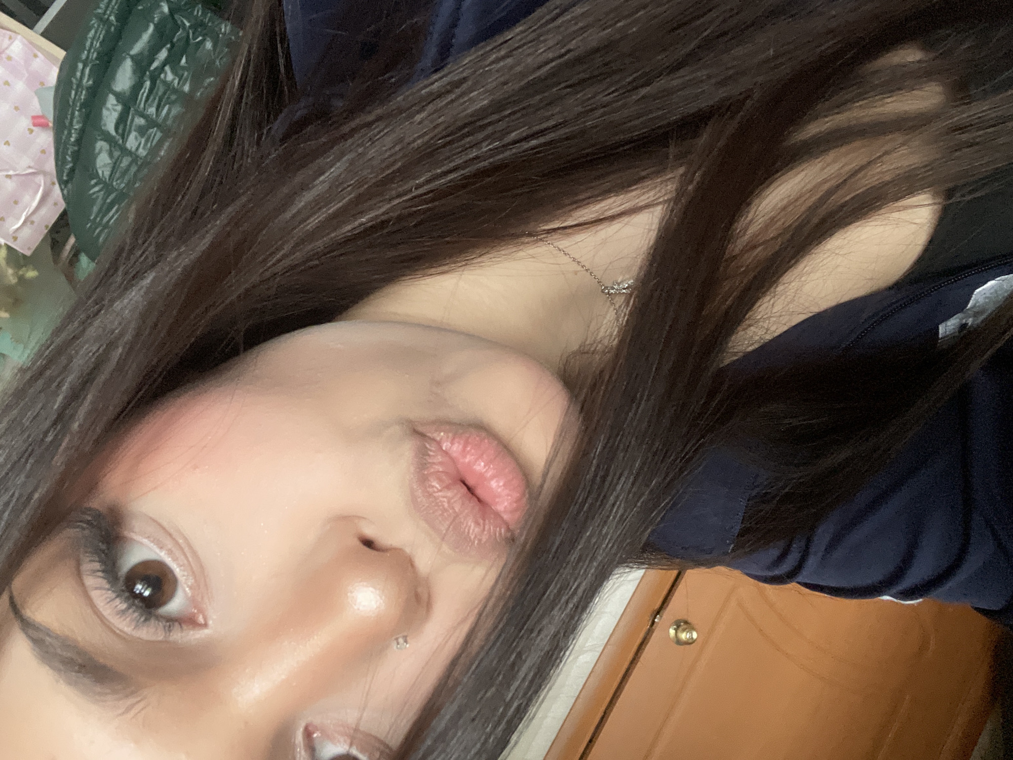 The image is a close-up, angled portrait of a person, rotated slightly counter-clockwise. The main subject is an individual with long, dark brown hair that frames the face and extends throughout the frame. The person has fair skin, dark brown eyes, and is looking directly at the camera. Makeup is visible, including blush on the cheeks, mascara on the eyelashes, and possibly lip gloss on the lips. A small, shiny piercing is present on the philtrum. Around the neck, a delicate silver chain necklace with a small, sparkling pendant is visible. The person is wearing a dark navy blue garment, possibly a jacket or sweatshirt.
The person's lips are pursed in a "kiss" or "pout" expression, suggesting a playful mood or pose for the photograph.
In the background, various indoor elements are visible. To the upper left, a dark green, reflective, possibly quilted or puffy fabric or object can be seen. Below it, a light-colored fabric with small pink heart patterns is partially visible. To the lower right, a warm brown wooden surface, likely a door, is present, featuring a circular brass-colored doorknob.
There are no discernible time-of-day indicators or weather conditions. No text is visible in the image. The overall setting appears to be an indoor room.