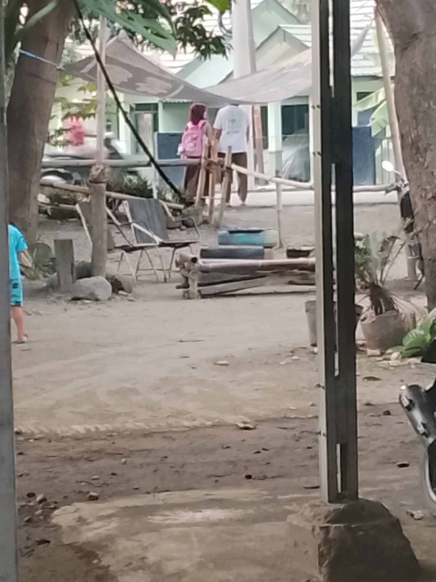 The image depicts an outdoor scene in Ujungrusi, Indonesia, with a sandy ground and some scattered debris. In the background, a building with a light-colored roof and greenish walls is visible, partially obscured by a canopy. Two individuals are present: one person, appearing to be a child, wears a pink hijab and a dark skirt, carrying a pink backpack. The other person, possibly an adult, is wearing a white t-shirt with a graphic on the back and dark pants. They are standing near a structure that resembles a covered walkway or an outdoor seating area. The foreground is dominated by a rough, unpaved surface with some rocks and a wooden structure. A tree trunk is prominently featured on the left side of the image, and on the far right, a motorcycle is partially visible. The overall impression is of a casual, outdoor environment.