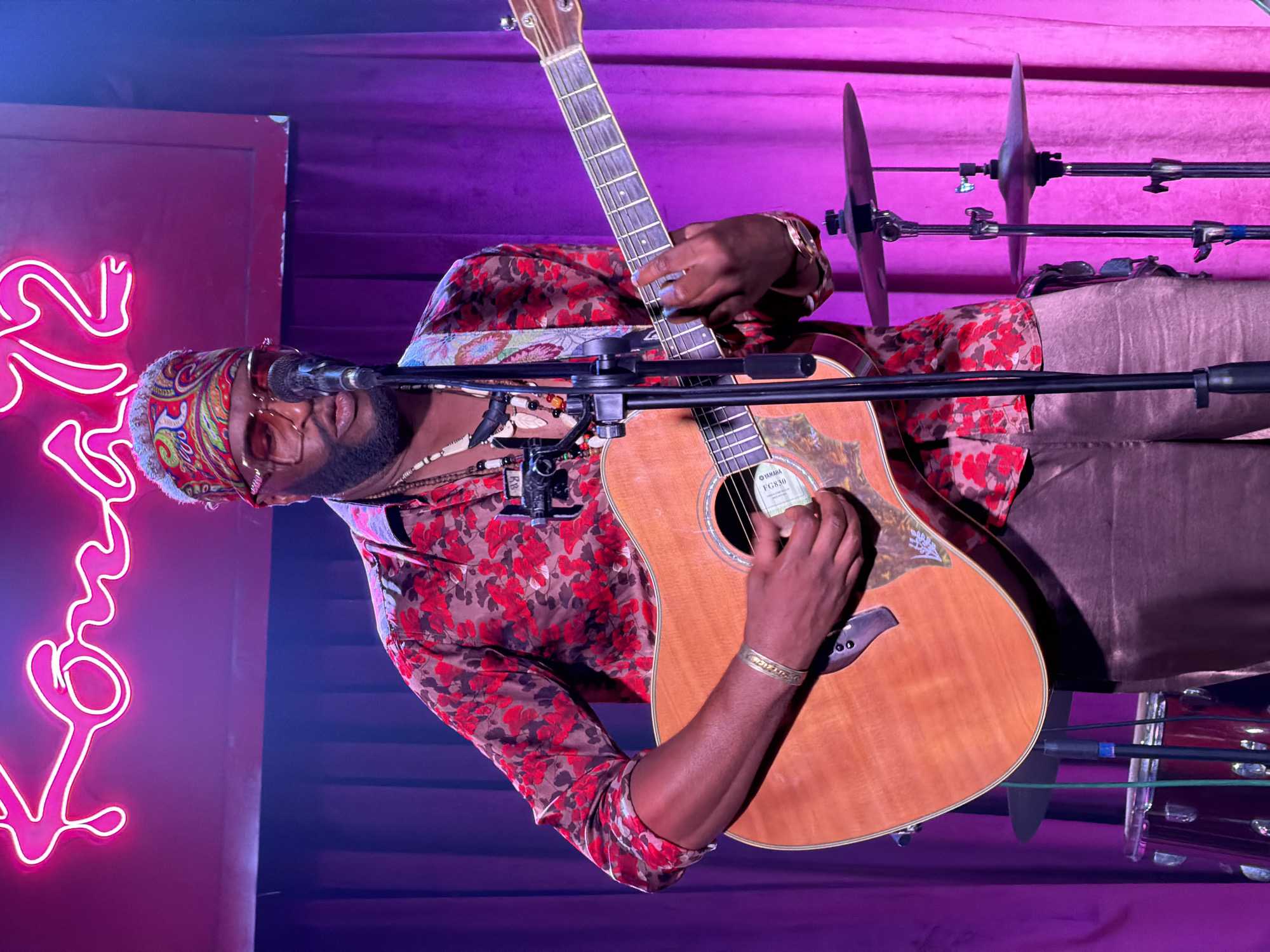 The image captures a male musician performing live indoors in Đà Nẵng, Vietnam. The central subject is a man with dark skin, a beard, and glasses, wearing a vibrant red and black floral-patterned long-sleeved shirt, tan trousers, and a colorful patterned headband. He also wears a beaded necklace and a golden bracelet on his right wrist, with his fingernails painted light blue.
He is actively playing a natural wood acoustic guitar, identified by a "YAMAHA FG830" label inside the soundhole. His left hand is on the fretboard, and his right hand is positioned near the soundhole, strumming the strings. A microphone on a stand is placed directly in front of him, suggesting he is also singing.
The setting appears to be a stage or performance area, likely an evening or night event due to the artificial lighting. The background features deep purple curtains, illuminated with strong purple and pink stage lights, creating an atmospheric glow. To the left, a bright pink neon sign is partially visible, displaying what appears to be the word "Live," likely part of a "Live Music" sign. To the right of the musician, parts of a drum kit, specifically cymbals on stands, are visible, indicating other instruments are part of the performance setup. The musician appears focused and immersed in his performance.