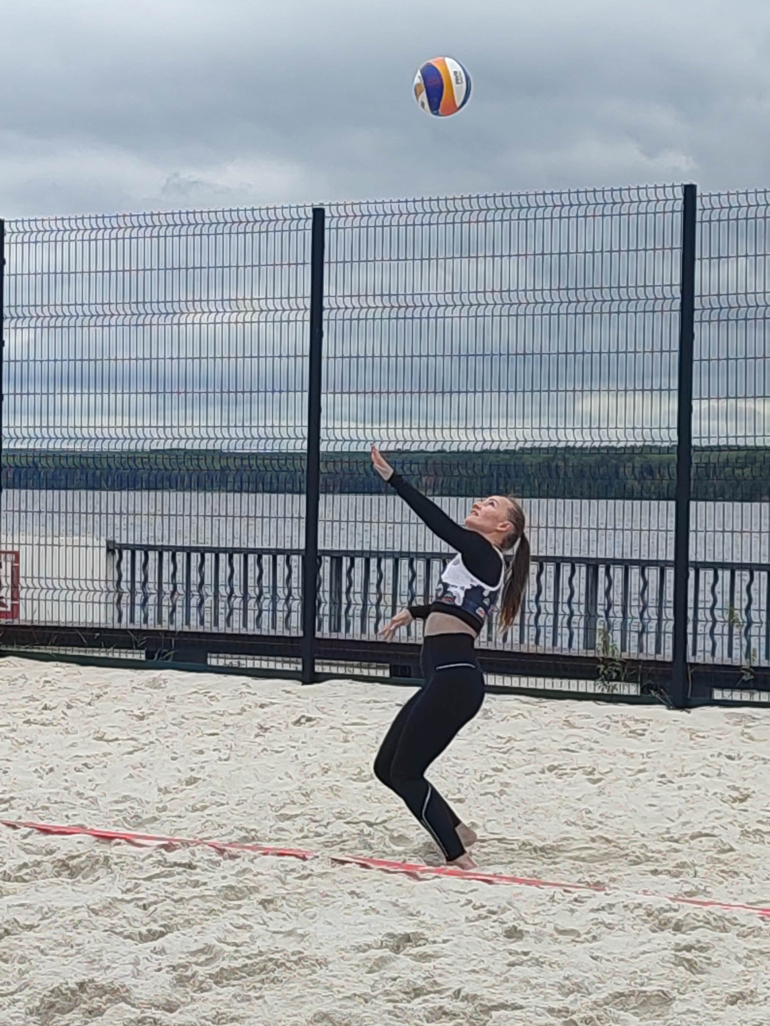 A young woman is playing volleyball on a sandy court. She is wearing black leggings and a black and white sports top. She is in the middle of serving the ball, with her arm extended and the ball in the air above her. The beach volleyball court has red boundary lines. In the background, there is a black metal fence, a body of water, and a line of trees. The sky is overcast and cloudy, suggesting it may be a cool or damp day. The image appears to be taken in daylight.