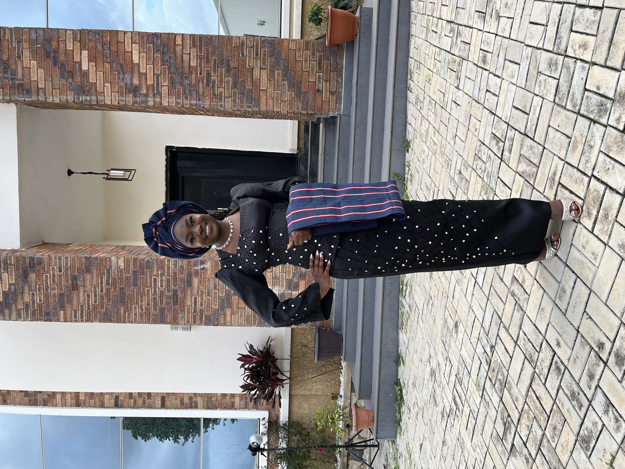 A young Black woman stands on a paved outdoor patio in front of a building with a brick facade and large glass windows. The woman is wearing a traditional black outfit adorned with white beads. Her head is wrapped in a blue and red patterned headscarf. She is holding a striped blue, white, and red fabric piece. She smiles directly at the camera with her left hand on her hip. To the left of the woman, a photographer's light is visible on a stand. Behind her, stone steps lead up to a dark doorway. The building appears to be modern and well-maintained, with potted plants visible in front of it. The overall setting suggests an outdoor event or photoshoot. The image is taken in Gura Top, Nigeria.