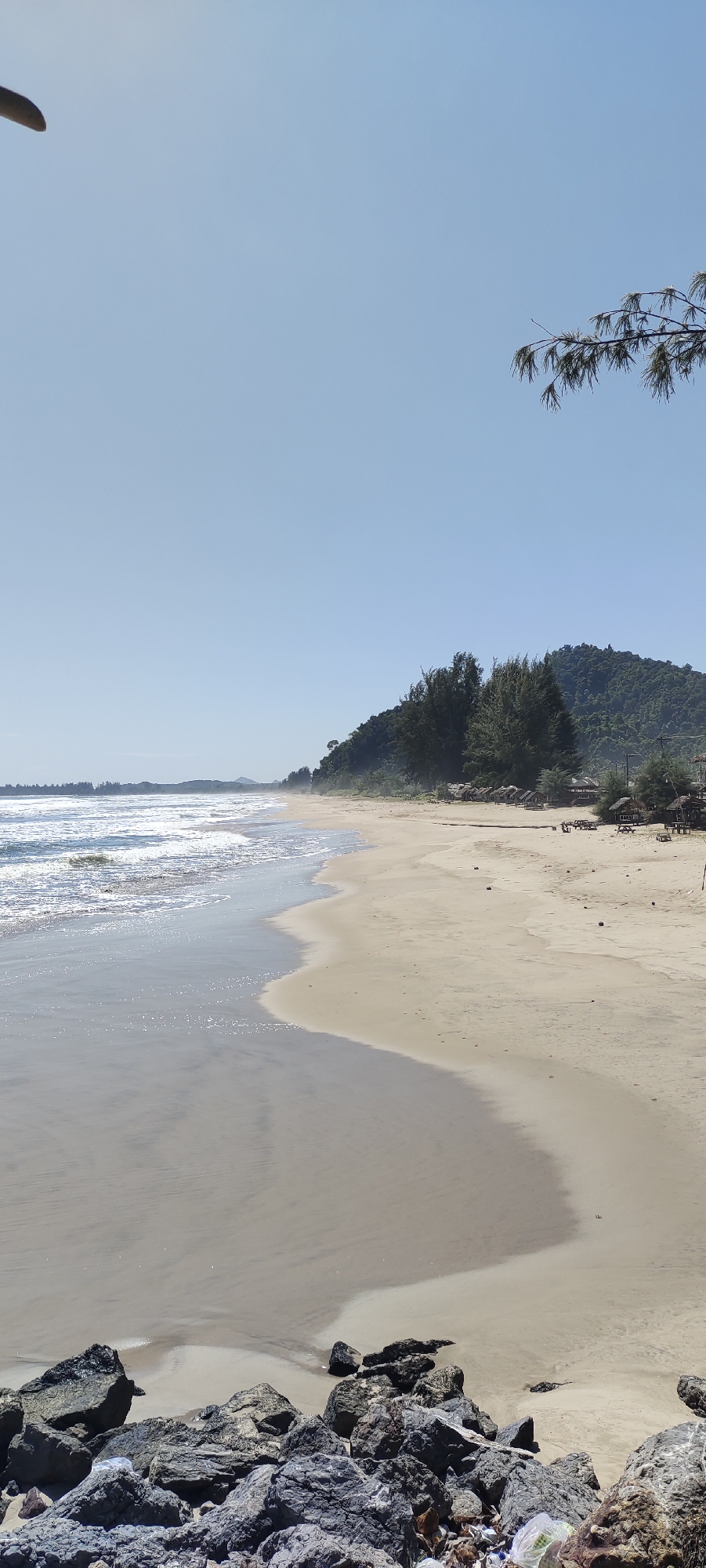 This image depicts a serene beach scene in Meudheun, Indonesia, under a clear blue sky. The foreground is dominated by a cluster of large, dark rocks, some appearing wet and glossy from the water. Beyond the rocks, the wet sand of the beach stretches out, meeting the gentle waves of the ocean. The ocean itself is a light blue, with white foam cresting on the waves.
The midground of the image features a wide expanse of dry, golden sand, leading towards a line of trees and a vegetated hillside. Scattered across the sand are several small, thatched-roof huts or cabanas, suggesting a resort or resting area. In the distance, the hillside rises, covered in dense green trees, with a faint haze or mist obscuring the very far background. The overall impression is one of a tranquil, natural coastal environment.