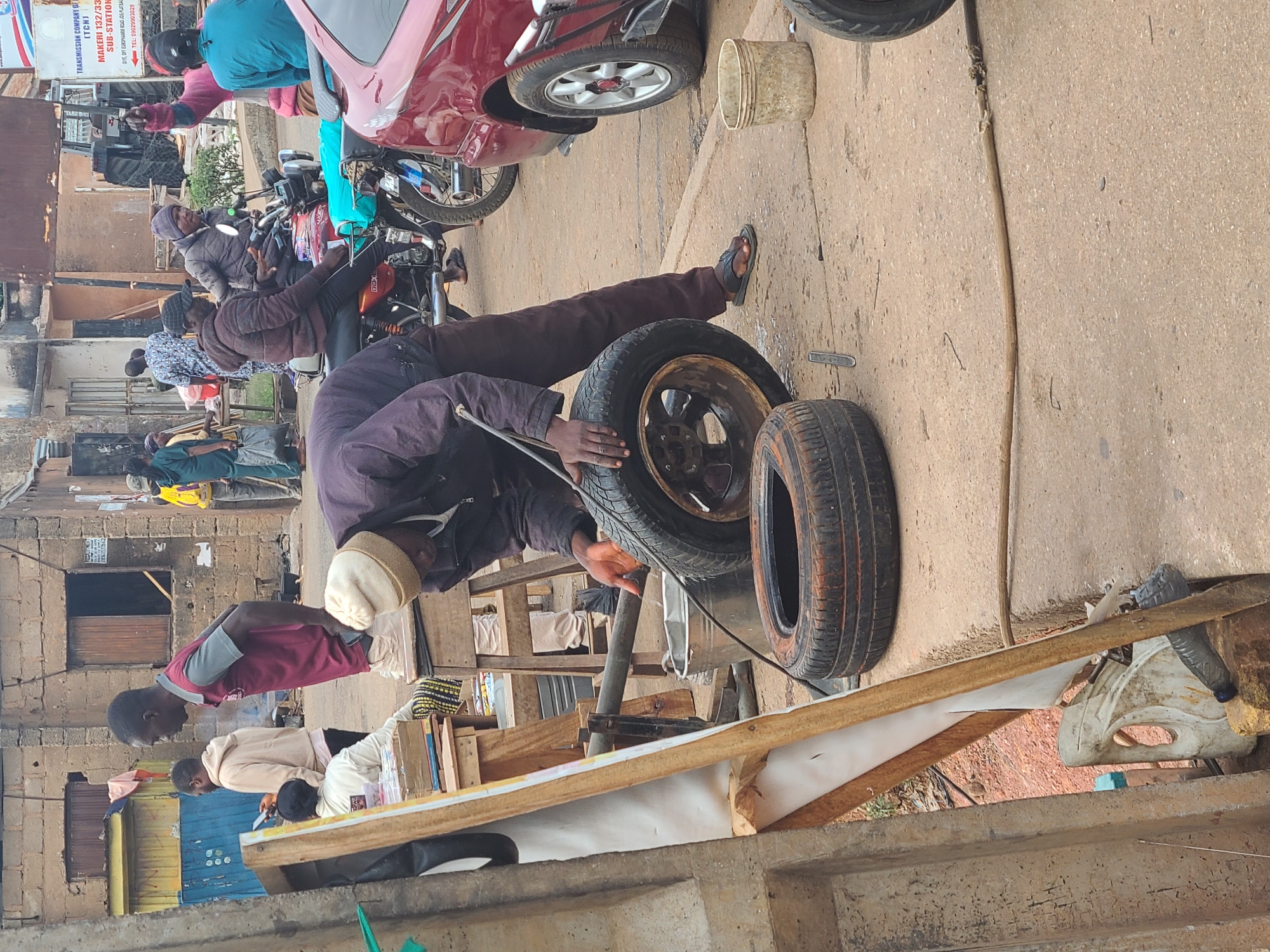 The image depicts an outdoor scene in what appears to be a bustling city, although the specific location is unknown. The foreground features a man working on a car tire. He is dressed in dark clothing and a light-colored hat. Two tires, one mounted on a rim, lie on the ground beside him. To the right is some sort of frame covered with wood. In the background are several parked cars, a few people standing and sitting, and buildings of varying heights. The sky is not visible, but the lighting suggests it is daytime. A sign mentioning "TRANSMISSION COMPANY OF (TCN) MAKERI 132/33 SUB-STATION" is attached to a building.