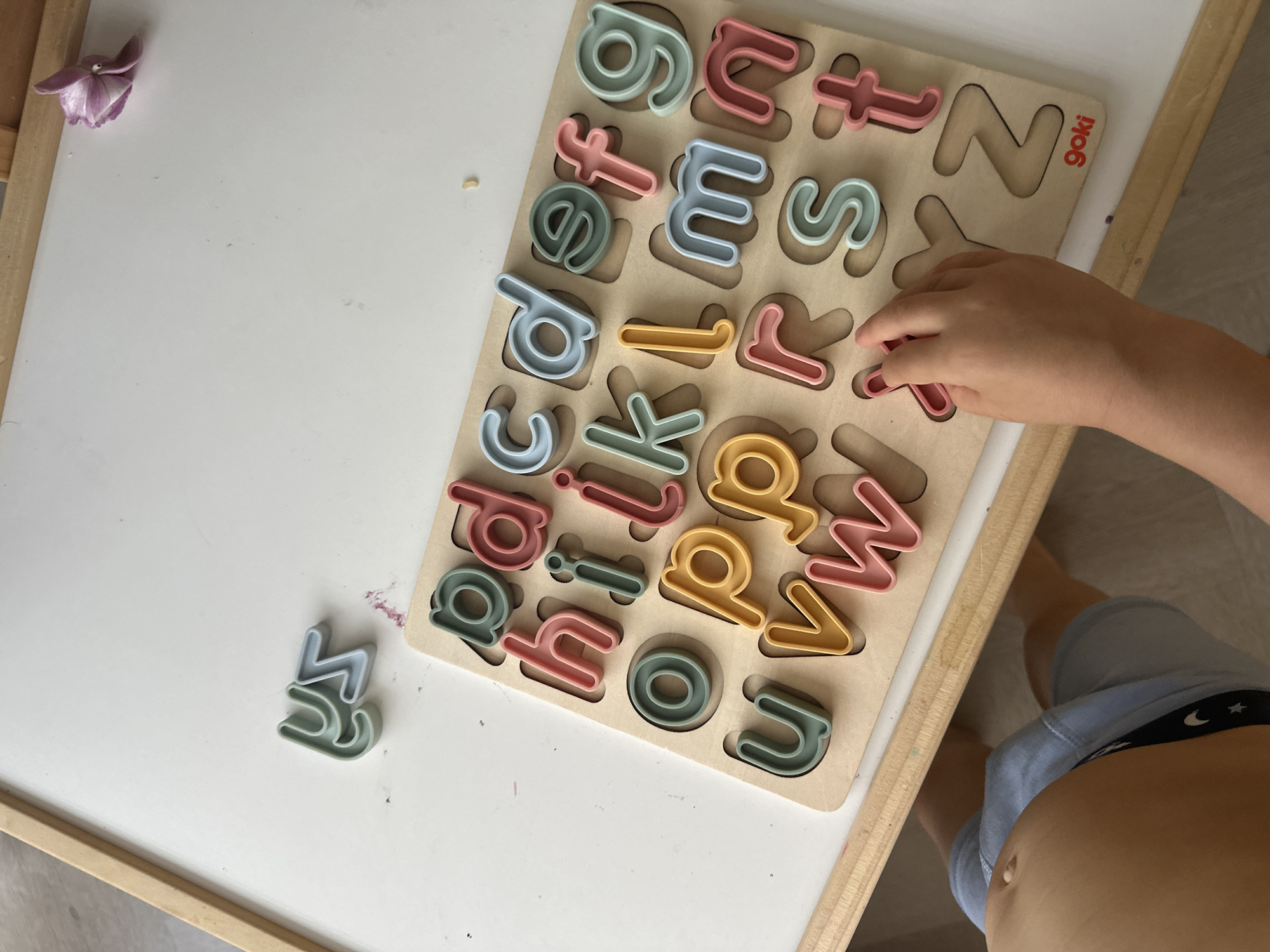 A child's hands are reaching for a wooden alphabet puzzle on a white surface, likely a table or easel. The puzzle contains pastel-colored lowercase letters arranged in alphabetical order, though some letters are out of place. The letters visible include 'a', 'b', 'c', 'd', 'e', 'f', 'g', 'h', 'i', 'j', 'k', 'l', 'm', 'n', 'o', 'p', 'q', 'r', 's', 't', 'u', 'v', 'w', 'x', 'y', and 'z'. The child is wearing blue shorts with a celestial pattern. The background shows the edge of a wooden easel or frame. A small purple flower is also visible on the white surface. The scene does not provide any information about the location in the city of Leeds, United Kingdom.