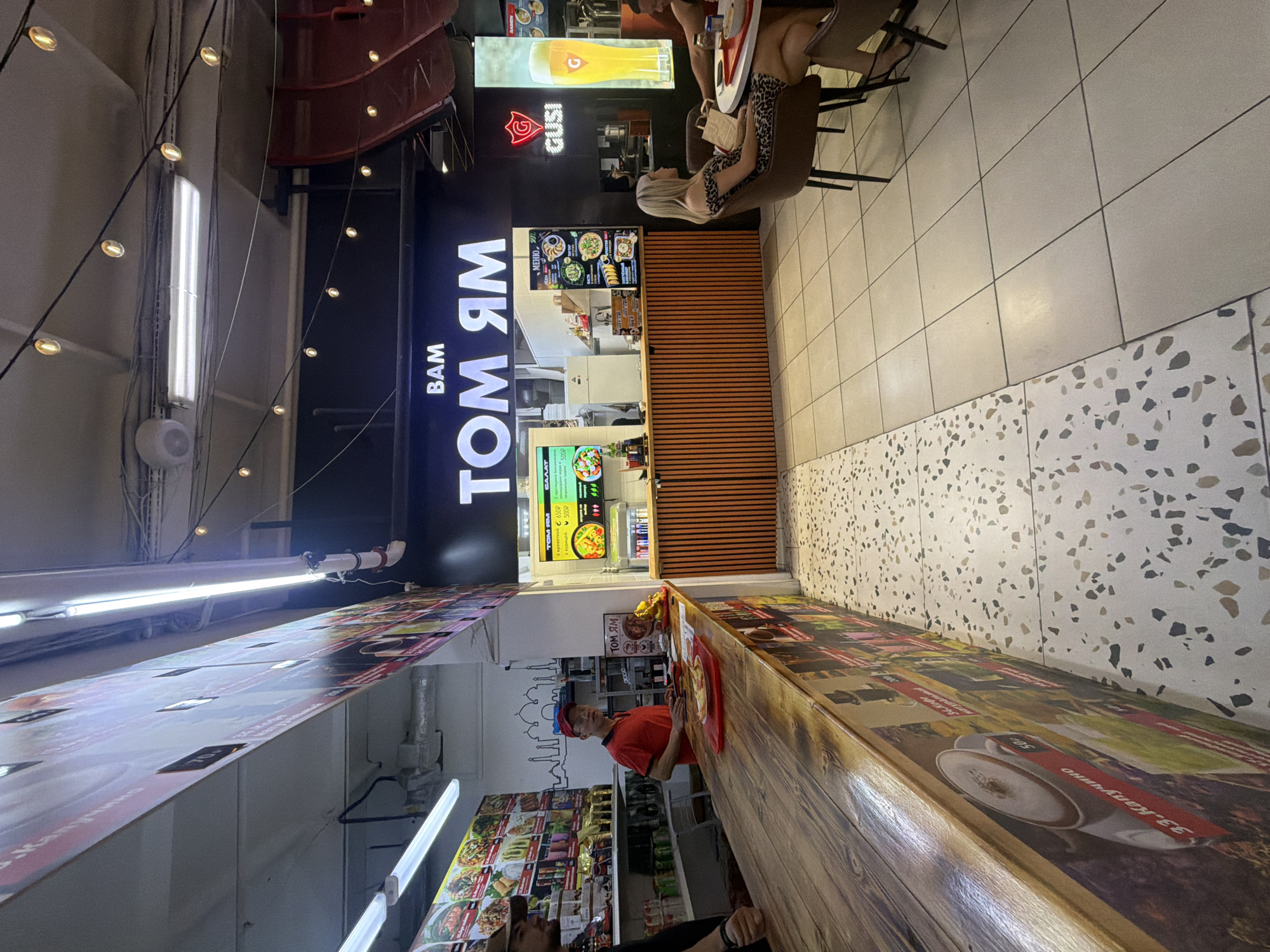 The image depicts an indoor scene, likely a food court or a restaurant establishment. The prominent signage reads "BAM TOM ЯМ", suggesting it is a Thai food restaurant. In the foreground, a wooden counter runs along the left side of the image, adorned with colorful graphics of food and drinks. A man in a red shirt and a red cap stands behind the counter, appearing to be an employee. A customer, partially visible on the left, is standing at the counter. On the right side, two individuals are seated at a round table. The restaurant features modern lighting, including fluorescent tubes and string lights. The floor is tiled with a speckled pattern in some areas and plain large tiles in others. The overall ambiance suggests a casual dining environment. There is no visible indication of the city of Novosibirsk, Russia, within the image.