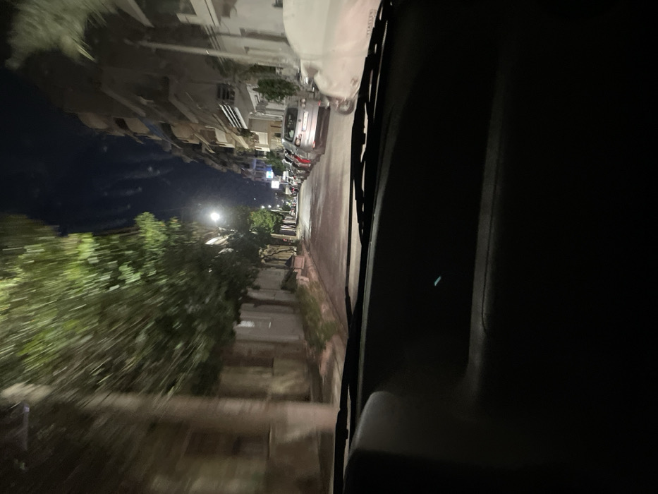 The image captures a nighttime street scene in Drapetsona, Greece, viewed from the interior of a moving vehicle. The perspective is from the front passenger side, looking out through the windshield.
In the foreground on the right, the dark interior of the car is visible, including a portion of the dashboard and the windshield frame. A small, faint blue-green light or reflection is present on the dark dashboard surface. Windshield wipers are discernible against the glass on the driver's side.
The midground reveals a street, which appears reflective, possibly due to wetness, extending into the distance. Parked cars line the right side of the road. Along the left side of the street, there is a sidewalk bordered by trees and bushes. These trees exhibit motion blur, indicating the vehicle from which the photo was taken is in motion. A bright streetlight illuminates the left side of the street and the foliage.
In the background, multi-story buildings, consistent with urban or residential architecture, line both sides of the street. The sky is dark, confirming the nighttime setting. On the rear of a white parked car, the faint lettering "FIAT" is visible. No people are seen. The scene evokes the quiet atmosphere of an urban area during the late hours.